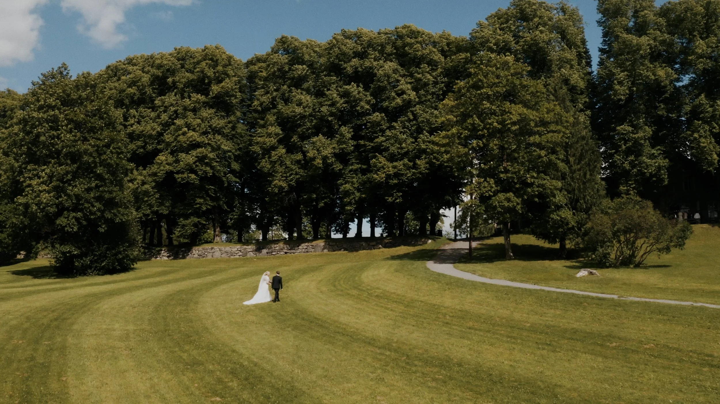 A bride and groom walk on a green grassy area near a winding path, with tall trees and a stone wall in the background on a sunny day.