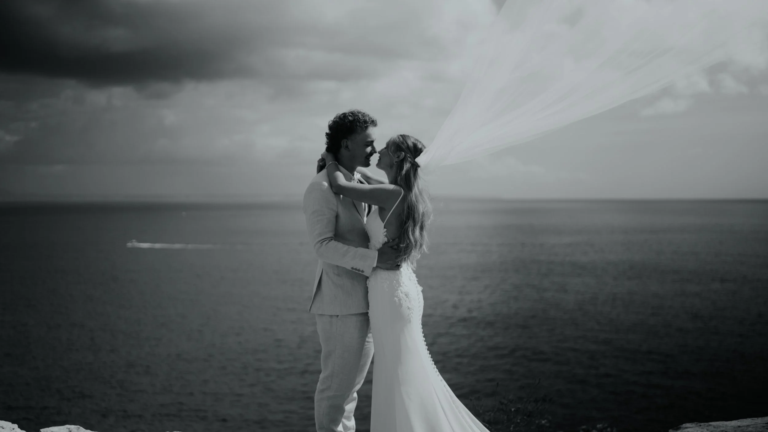 A black-and-white photo of a bride and groom embracing by the sea, with stormy clouds overhead and her veil flowing in the wind.