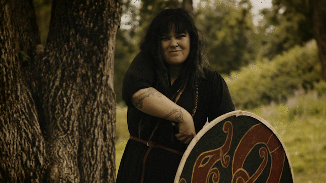 A woman with black hair and tattoos, wearing a black dress, standing outdoors next to a tree, with a decorative shield or round object in front of her.