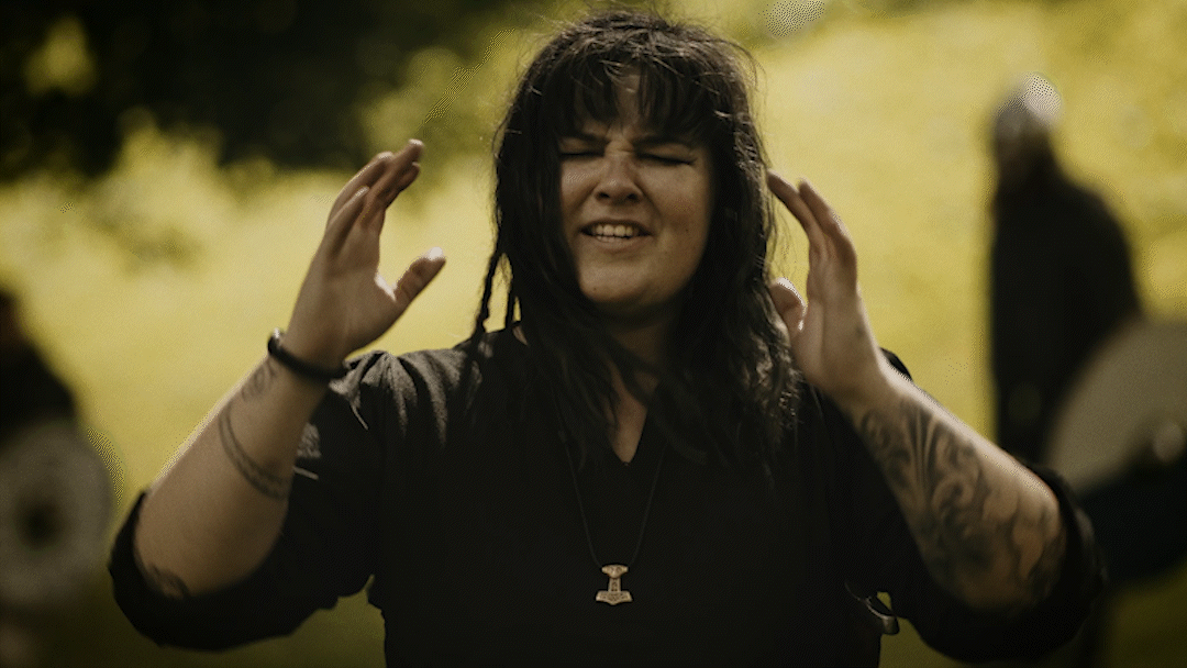 A woman with black, wavy hair and tattoos on her arms is outside with a blurred background, wearing a black shirt and a necklace with a pendant, making a gesture with her hands near her head and smiling.