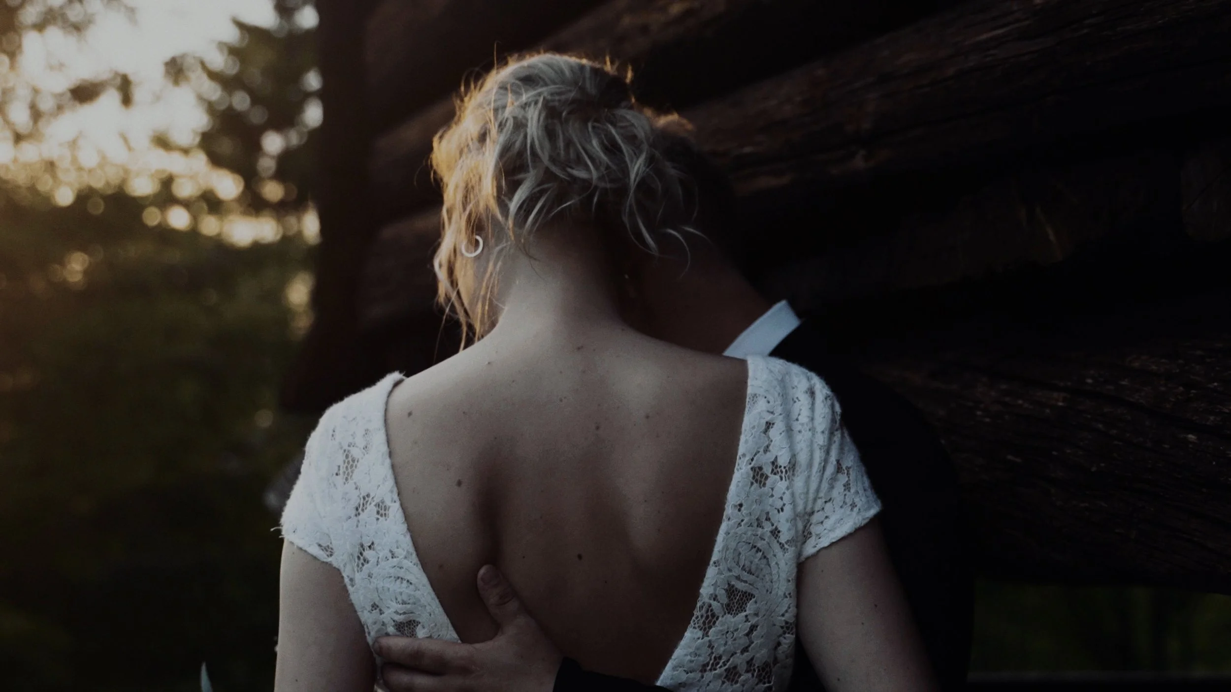 A woman with short blonde hair wearing a white lace dress and a man in a black suit, embracing outdoors near a large tree during sunset.