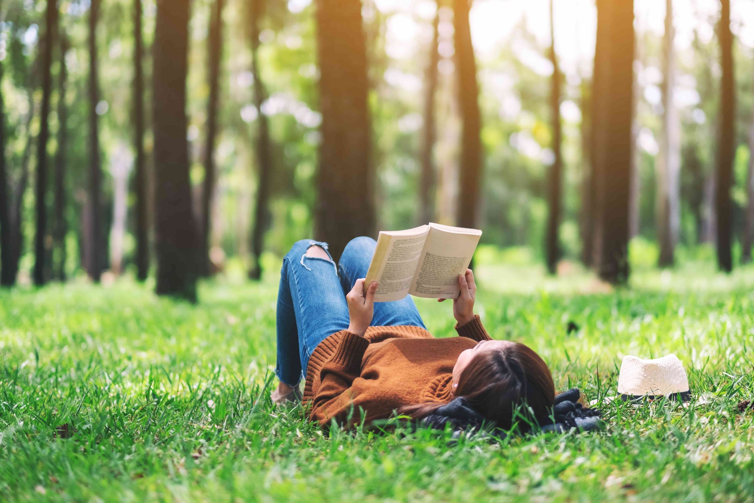 Person Lying Down On The Grass Surrounded By Trees Reading A Book