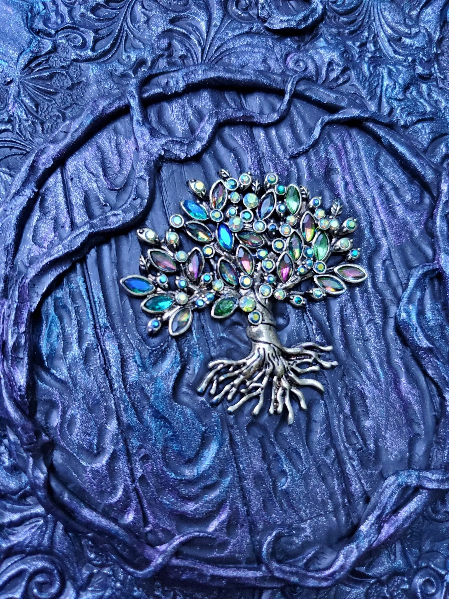 Decorative metallic ornament of a branching tree with colorful iridescent gemstones on a textured, ornate purple background.