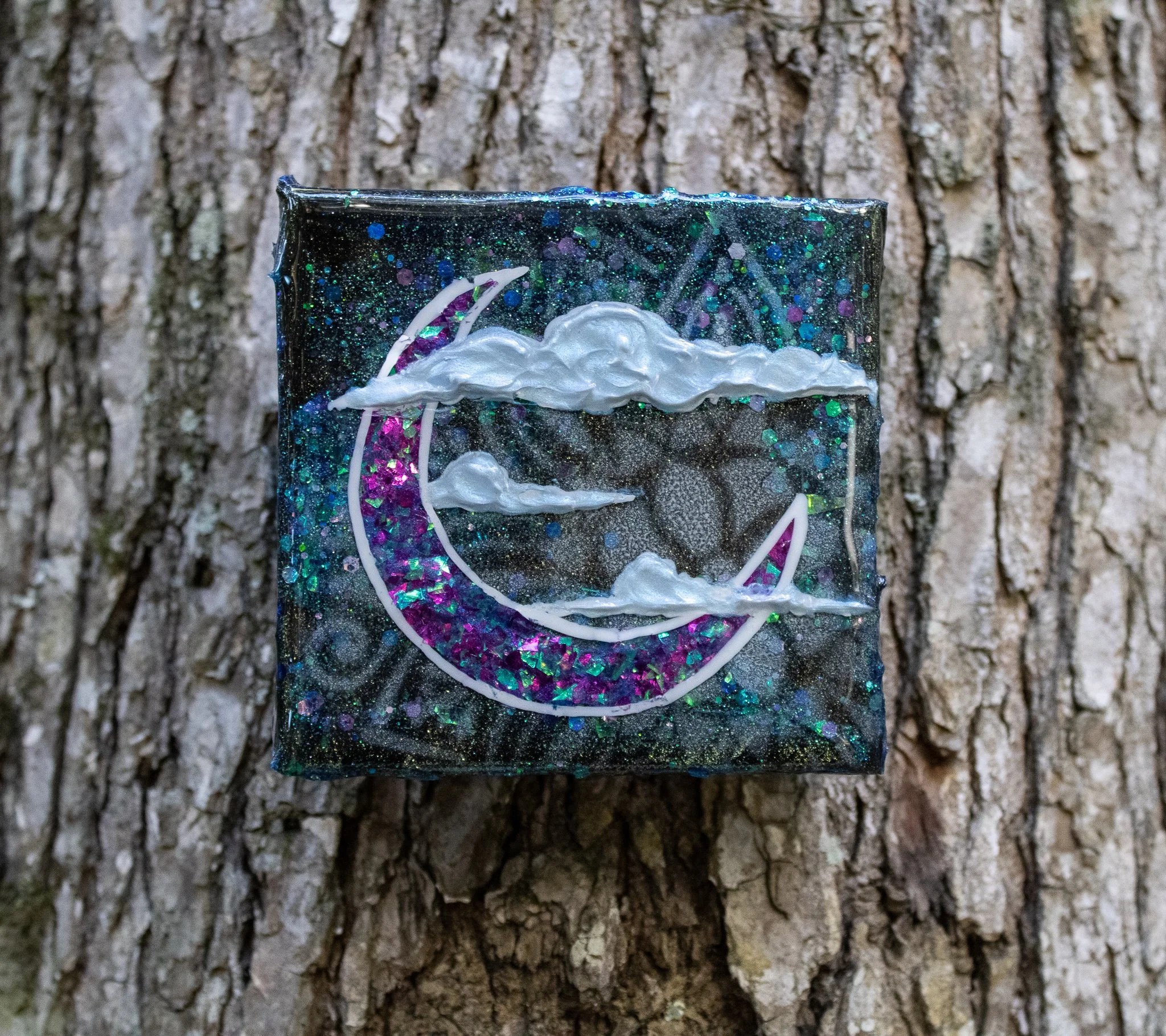 A small decorative box with a glittery galaxy theme, featuring a crescent moon and clouds against a tree bark background.