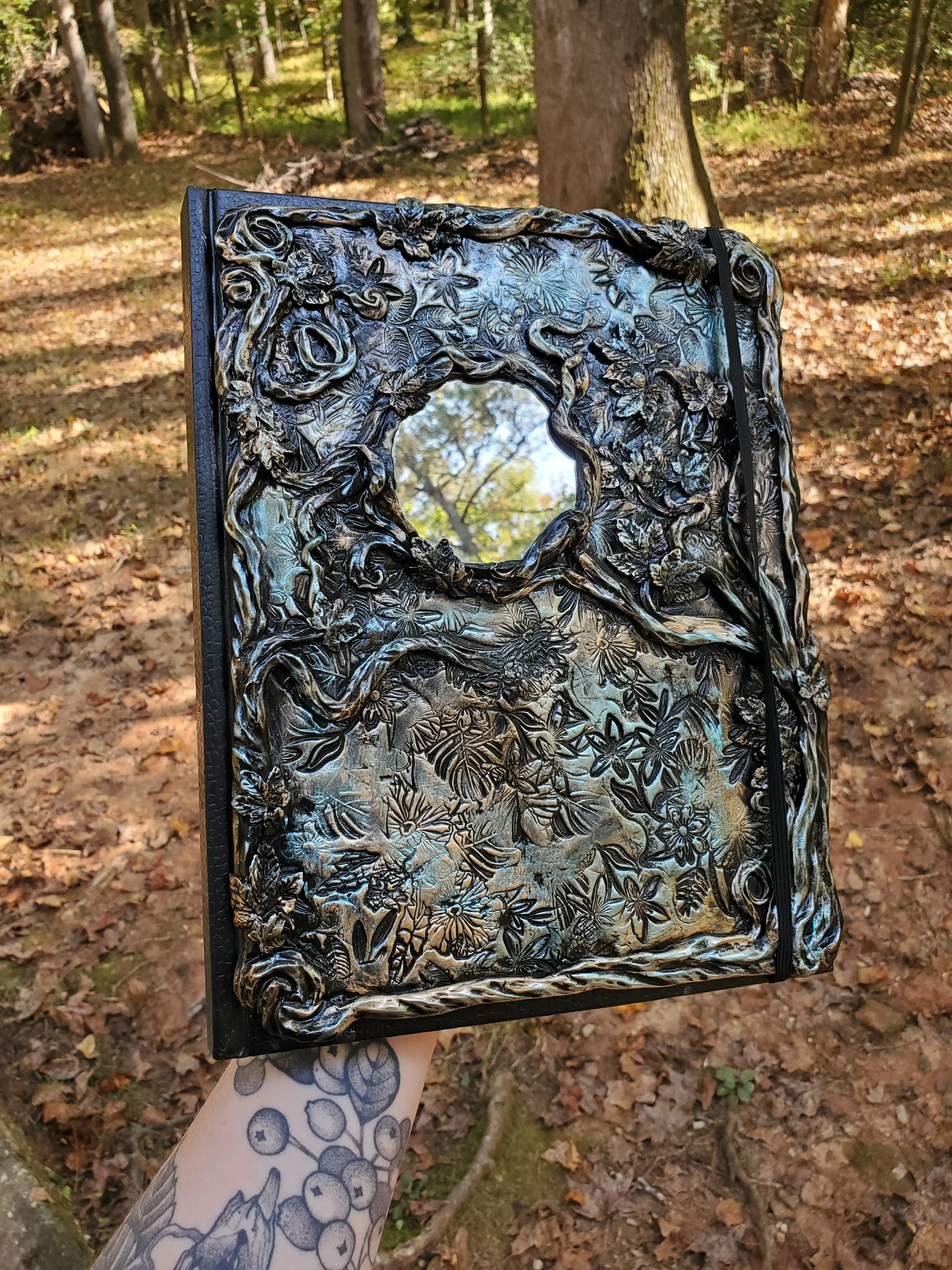 A decorative black leather-bound journal with intricate floral and vine embossing, held outdoors in a wooded area.