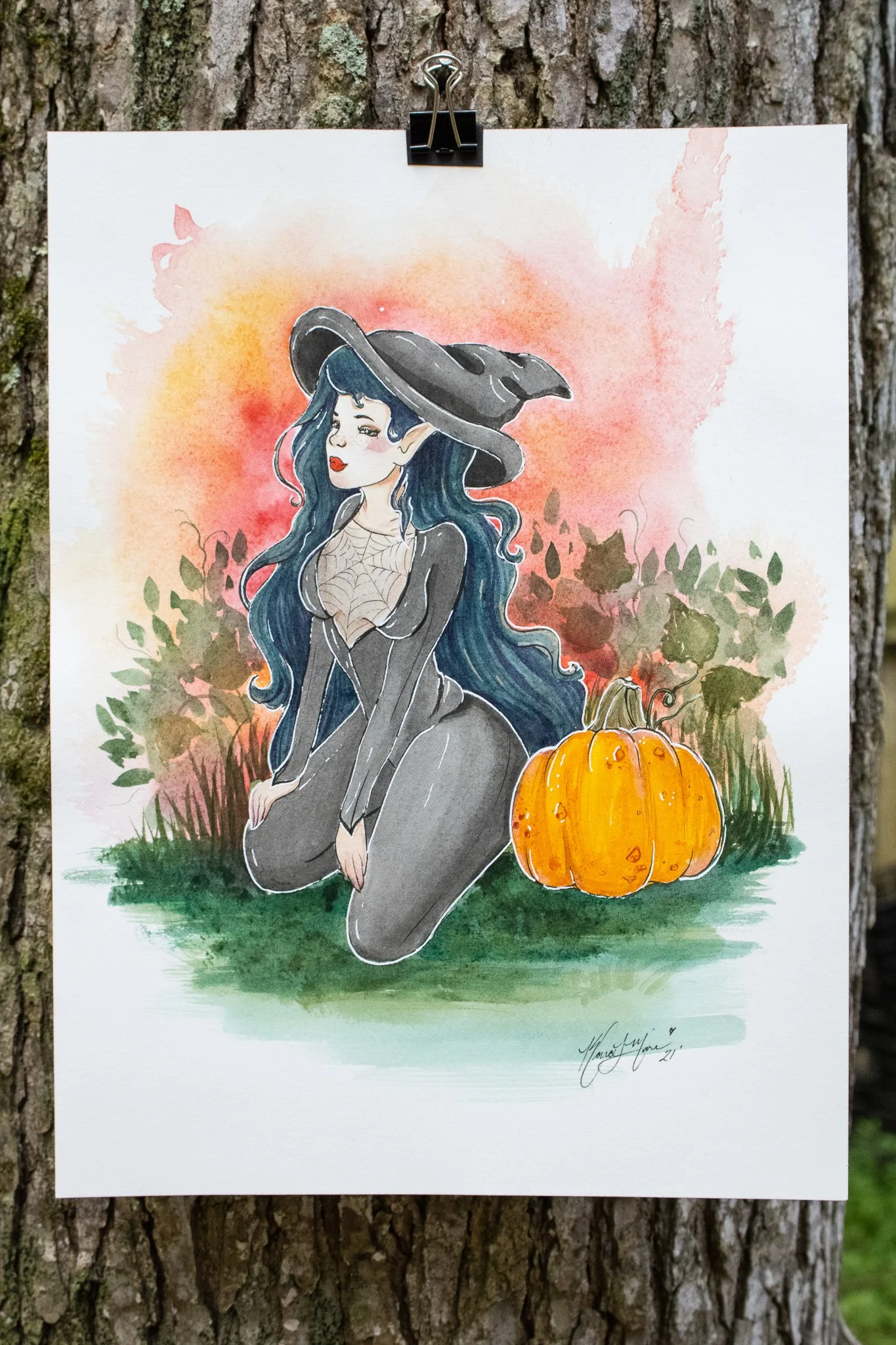 Watercolor painting of a witch with long blue hair, wearing a black hat and black dress, kneeling next to a pumpkin, against a pink and orange background with green foliage.