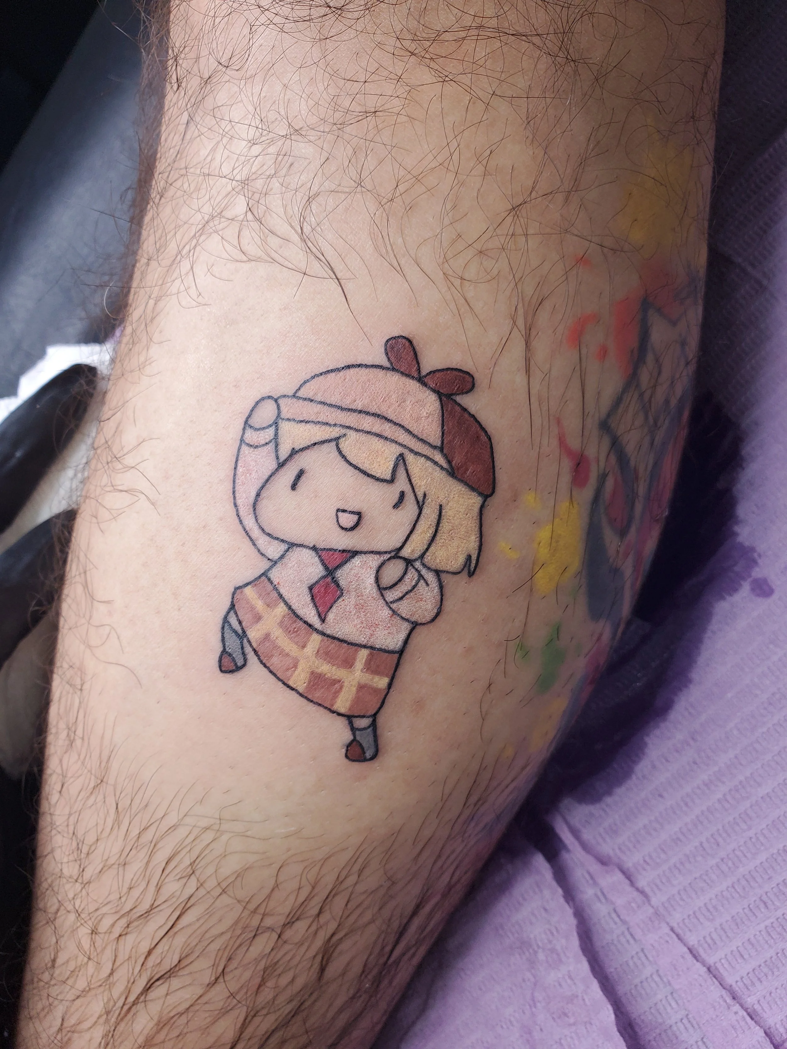 A colorful cartoon-style tattoo of a cheerful girl with blonde hair, wearing a beret, bookshelf skirt, red tie, and waving. Amelia Tattoo