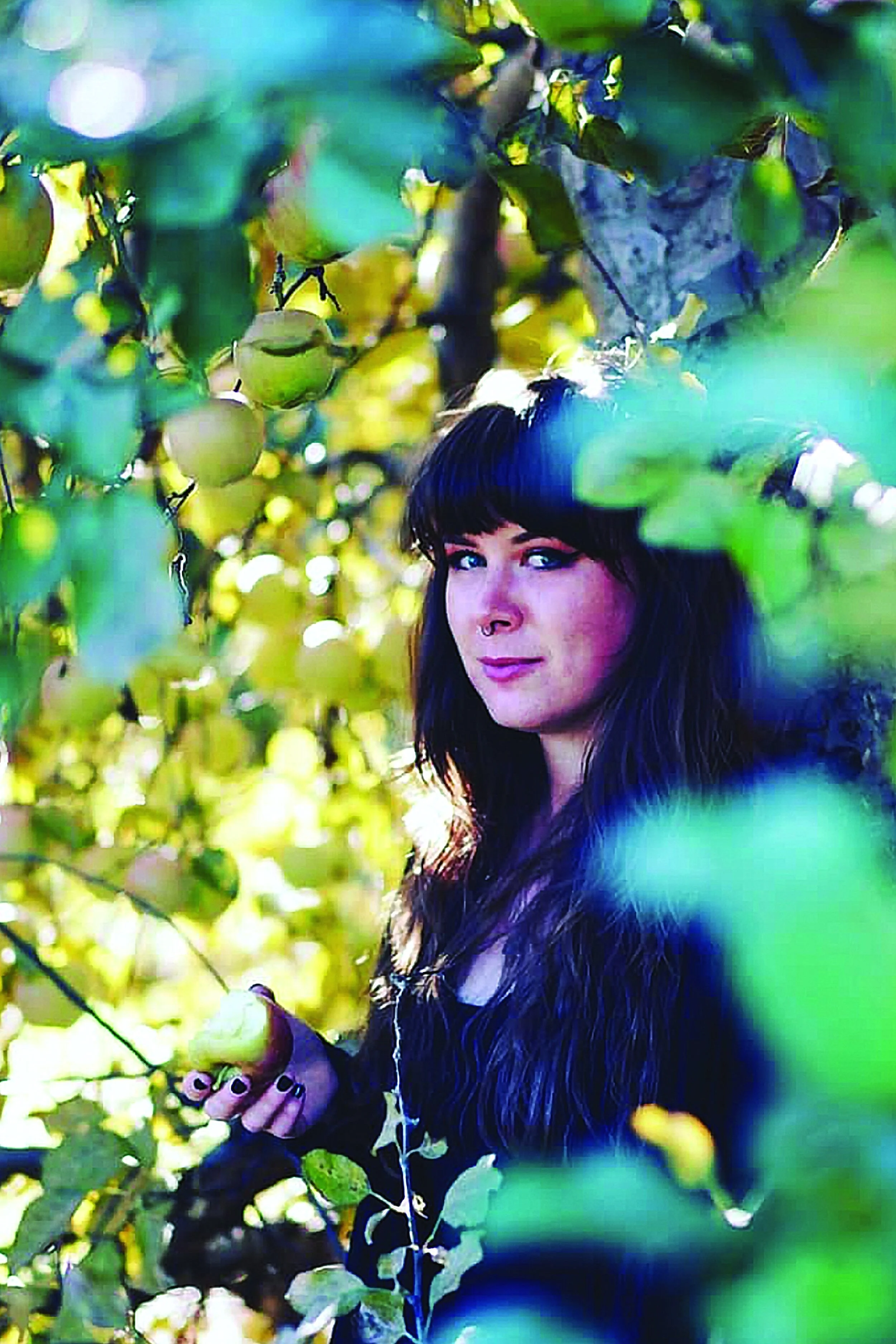 A woman with long dark hair, wearing dark clothing, stands amidst green leaves and fruit, holding a leaf in her hand, with sunlight filtering through the foliage.