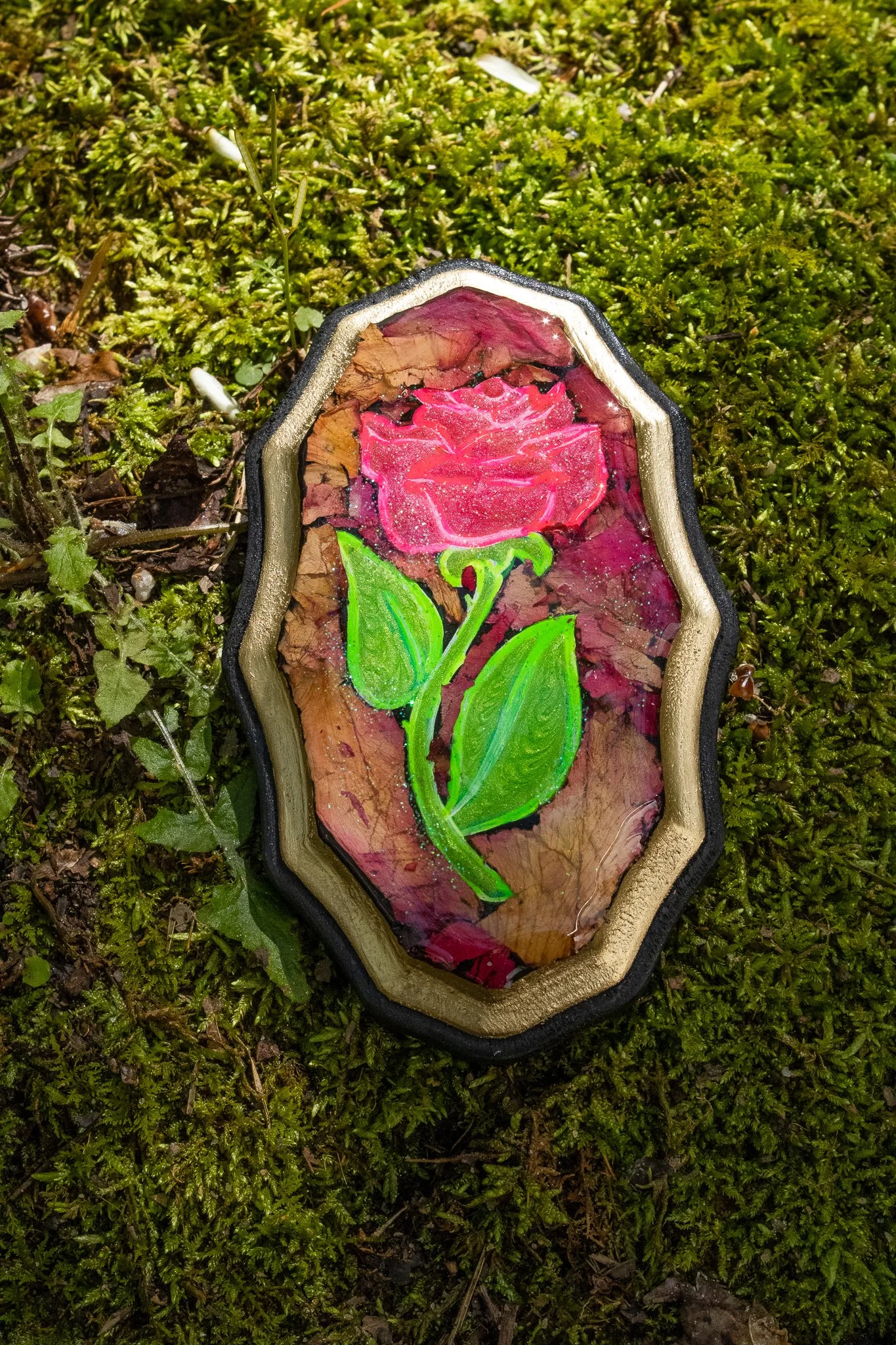 A decorative object with a painted pink rose and green leaves on a bark background, encased in an ornate border, lying on moss.