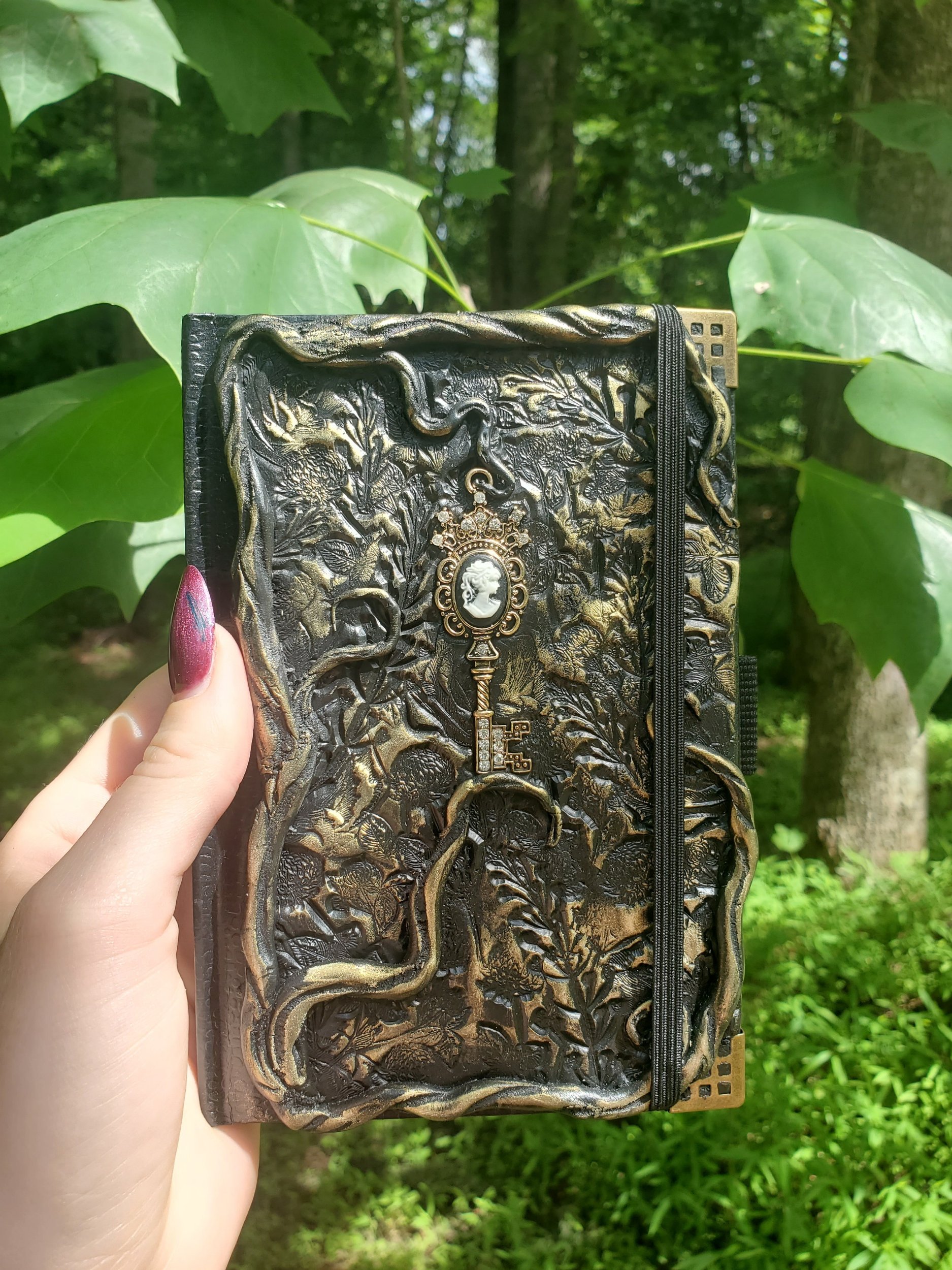 A person holding a decorative black and gold notebook cover with floral and tree designs, and a miniature key brooch or pendant in the center, against a background of green leaves and tree trunks.