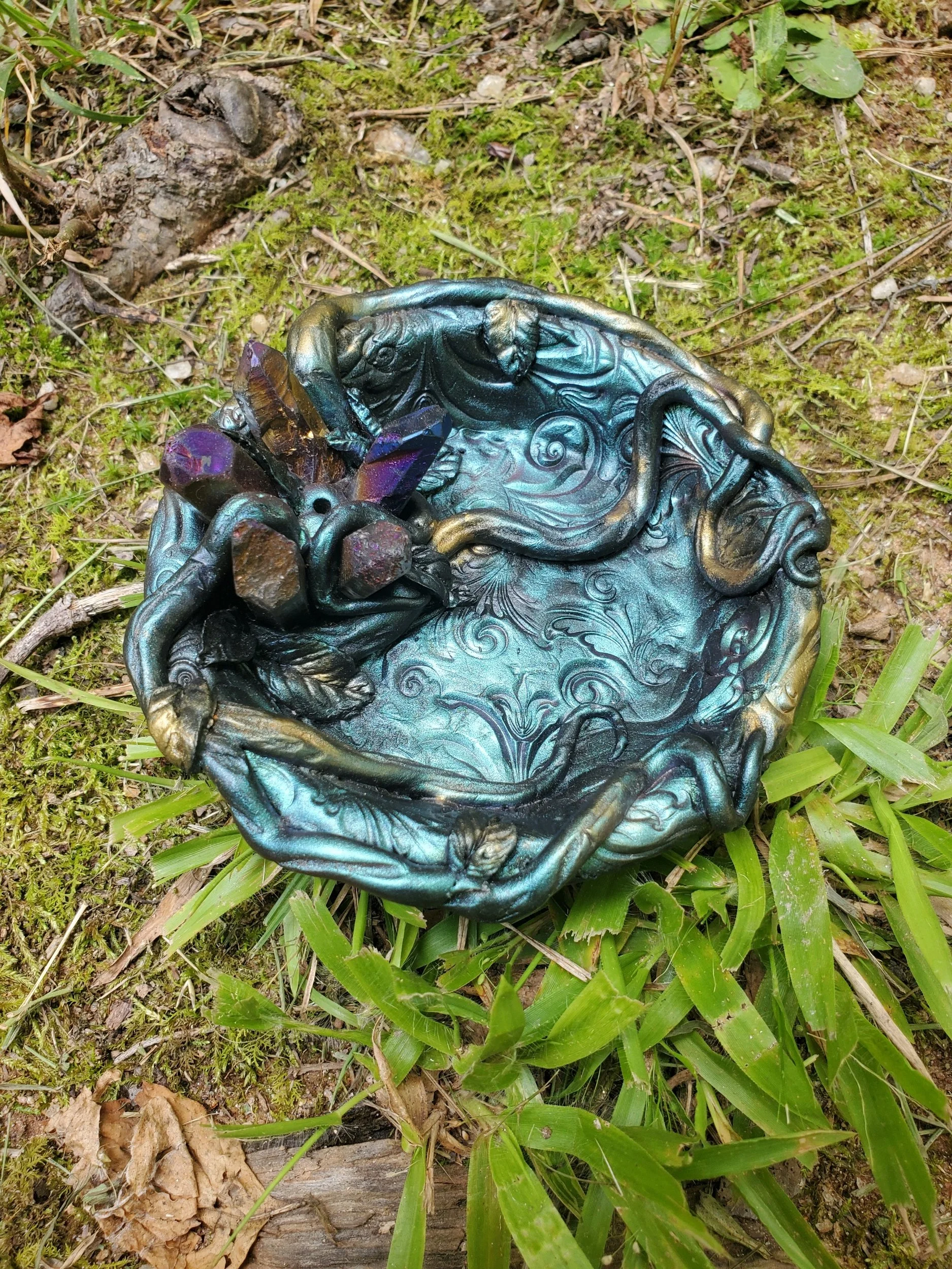 Decorative metallic dish with purple crystals, resting on grass and surrounded by moss and twigs.