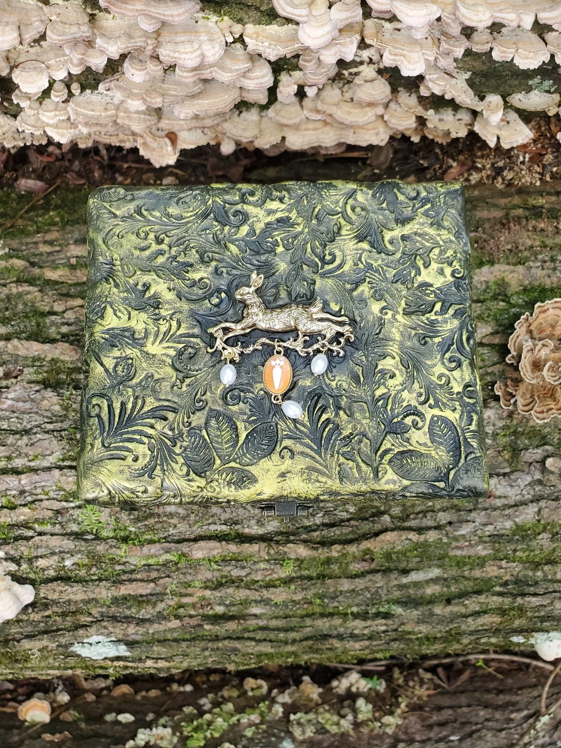 A decorative box with intricate engraved patterns and a small rabbit figure on the front, hanging on a tree trunk in a forest, surrounded by mushrooms and moss.