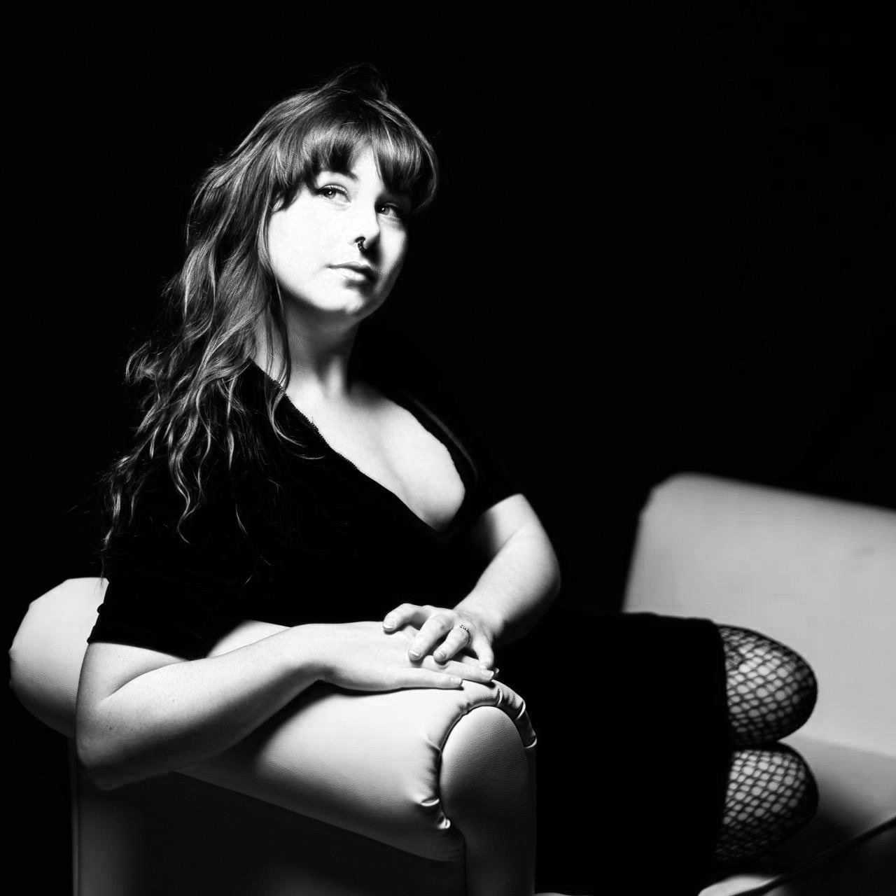 Black and white portrait of a woman sitting on a white chair with dark background, wearing fishnet stockings and a black top.