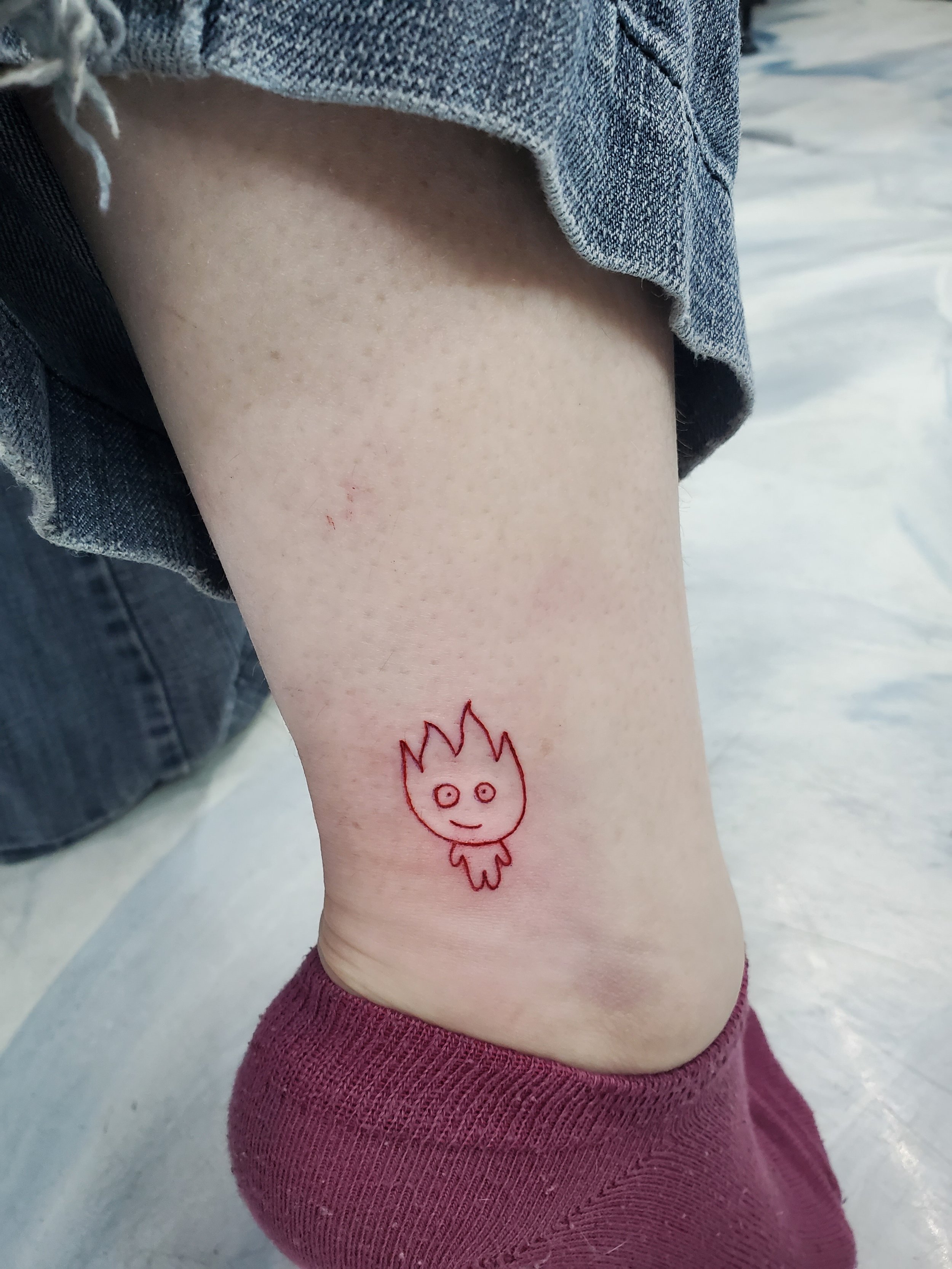 A small tattoo of a cartoon-like fire character with a smiling face on the person's ankle, near the sock line.