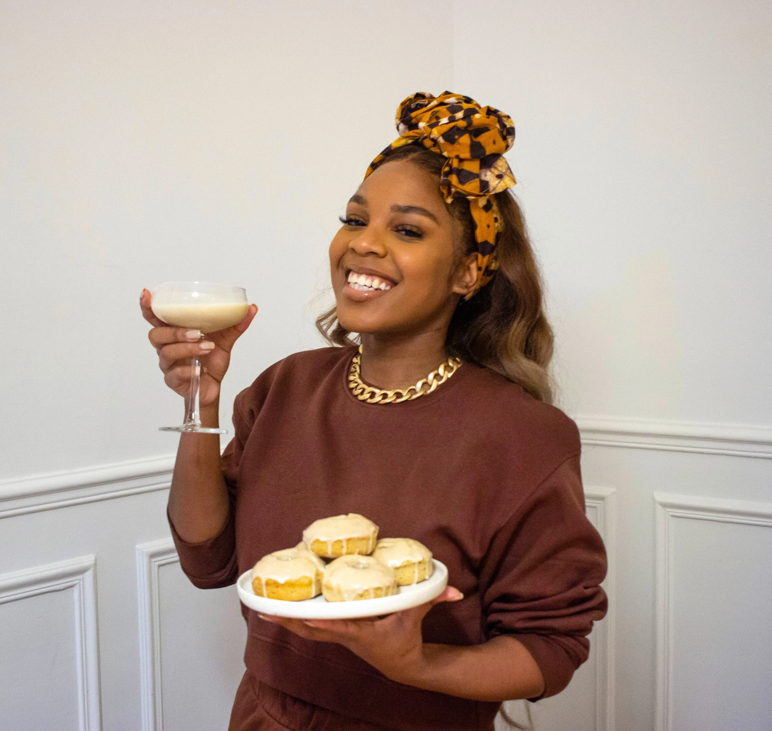 A woman smiling while holding a plate of cookies in one hand and a cocktail glass in the other, wearing a brown sweatshirt, a gold chain necklace, and a yellow and black headscarf.