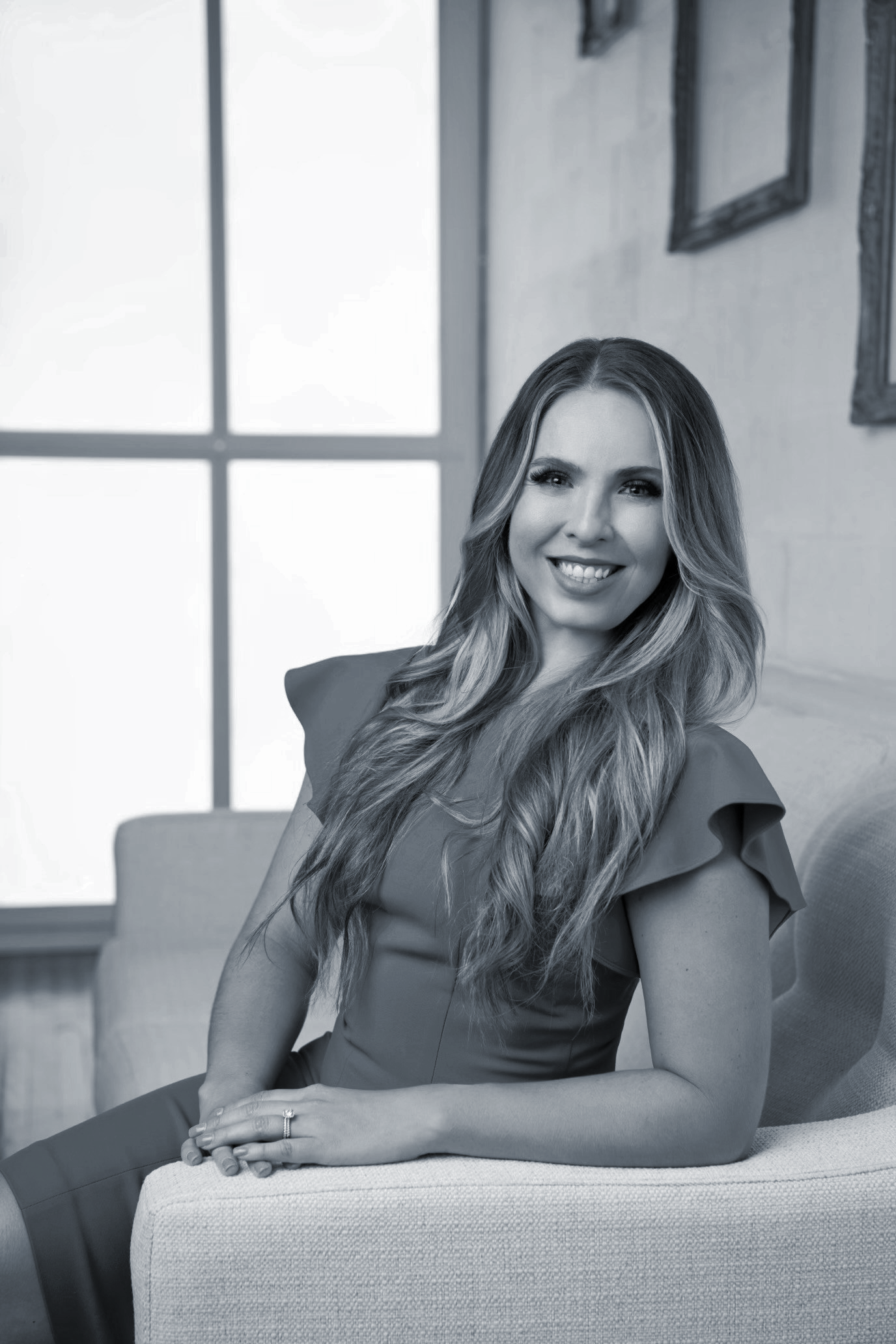 Black and white portrait of a smiling woman with long wavy hair, sitting on a sofa in a bright room with large window