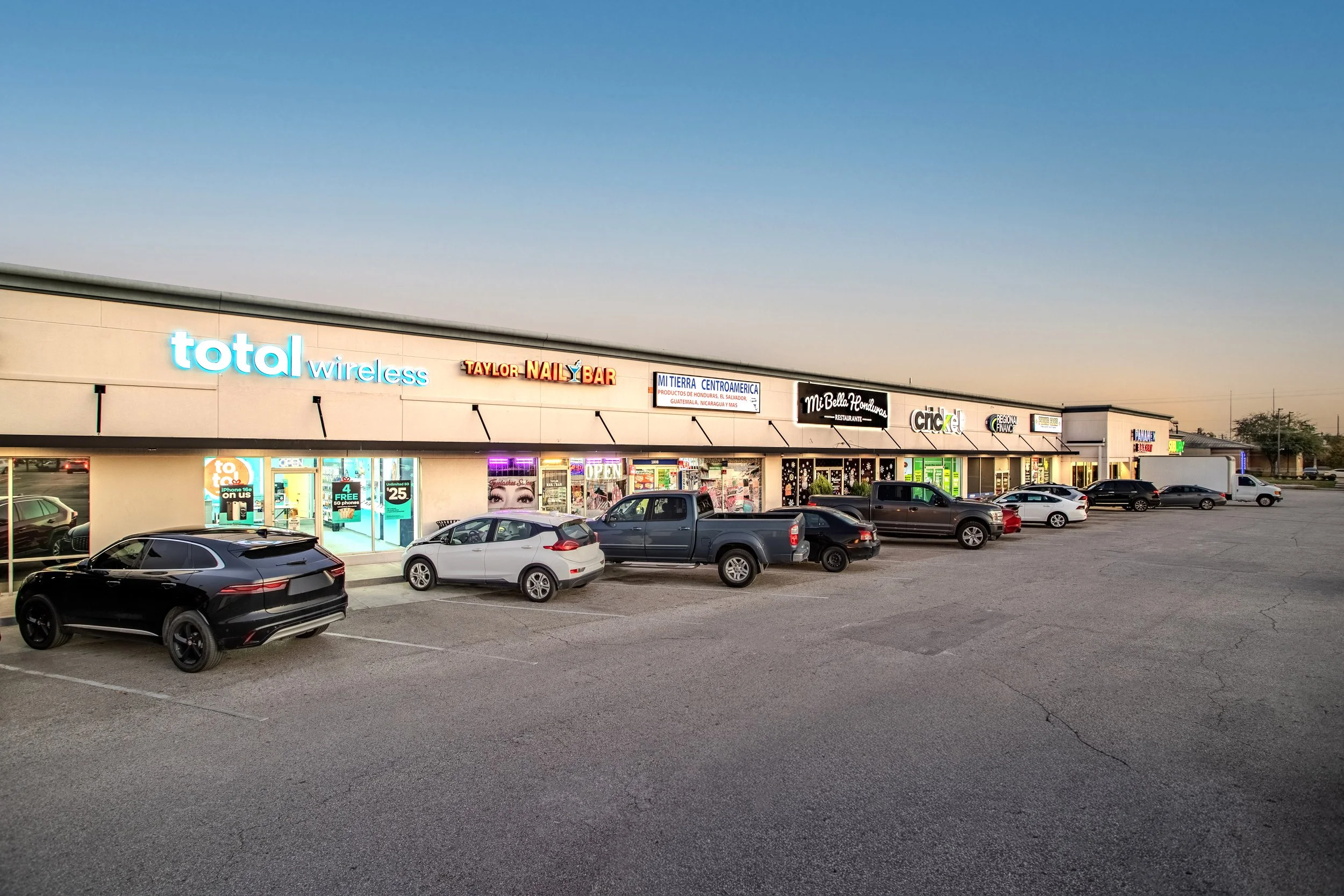 Shopping strip mall at dusk with various storefronts and parked cars in front.