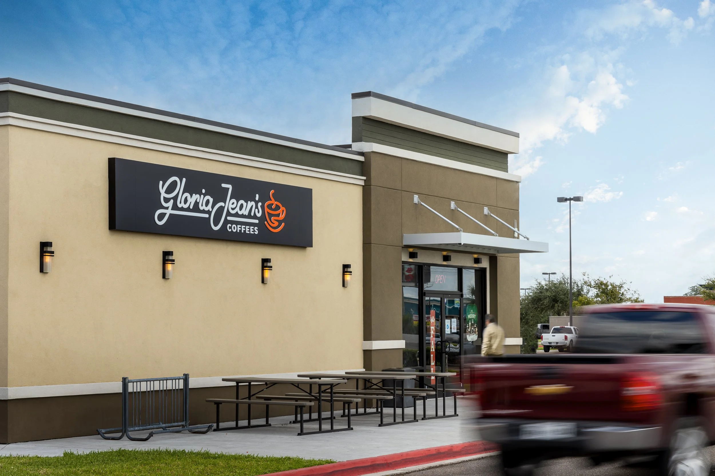 Exterior of Gloria Jean's Coffees shop with a sign, outdoor seating area with tables and benches, and a person near the door. A red vehicle is passing by.
