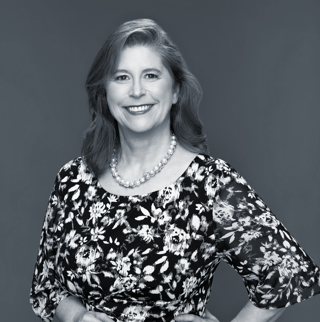 A woman wearing glasses and a sleeveless top, standing outdoors near a wooden wall with trees in the background.