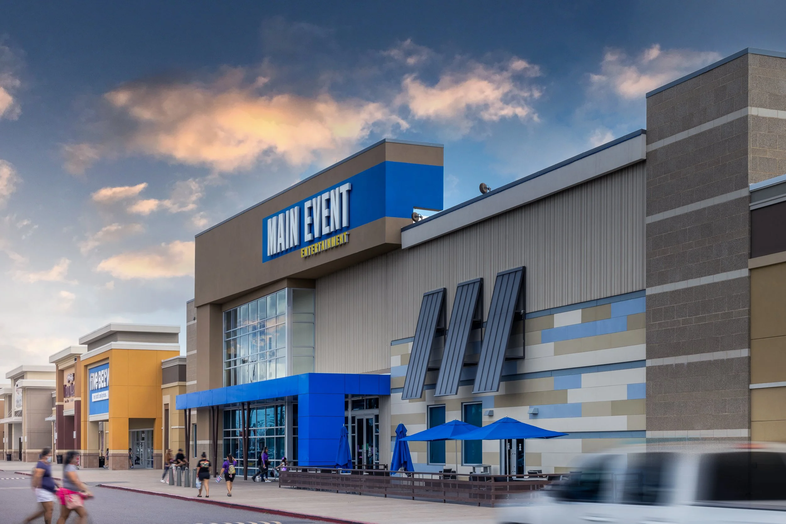 View of Main Event entertainment center with a prominent sign, a blue entrance canopy, and a parking lot with people walking and a moving car in the foreground.
