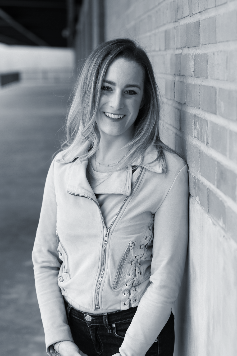 A woman with long hair smiling and leaning against a brick wall in an outdoor setting.