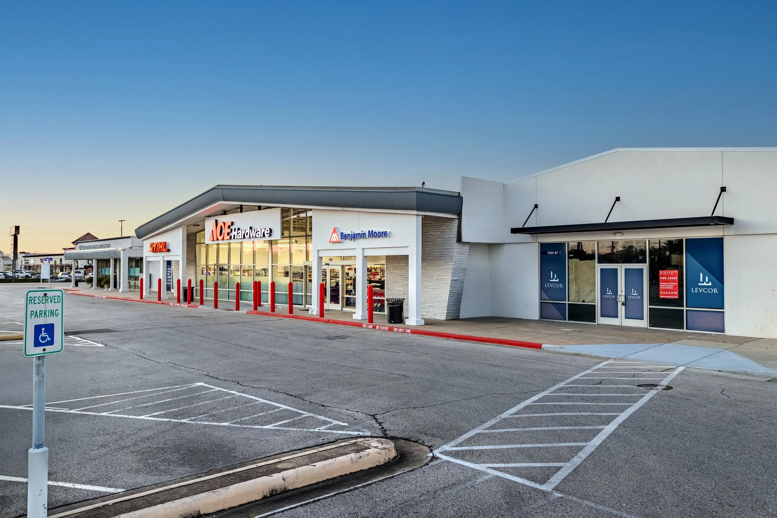 Empty parking lot outside a retail store with reserved handicap parking sign, storefront displays for Ace Hardware, Benjamin Moore, and Levcorr, with sunset sky overhead.