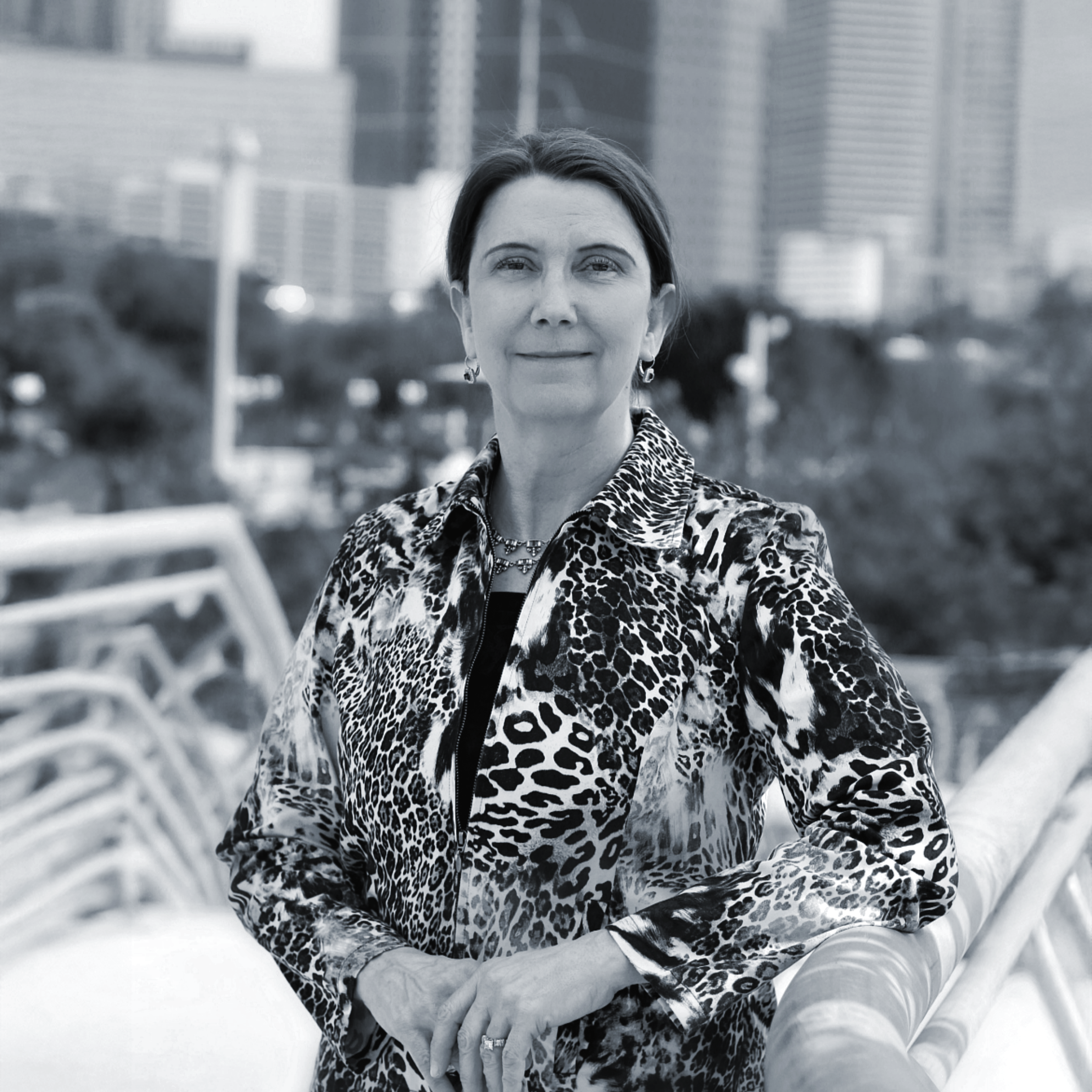 A woman with short dark hair, wearing earrings, a necklace, and a leopard print jacket, standing outdoors with a cityscape in the background.