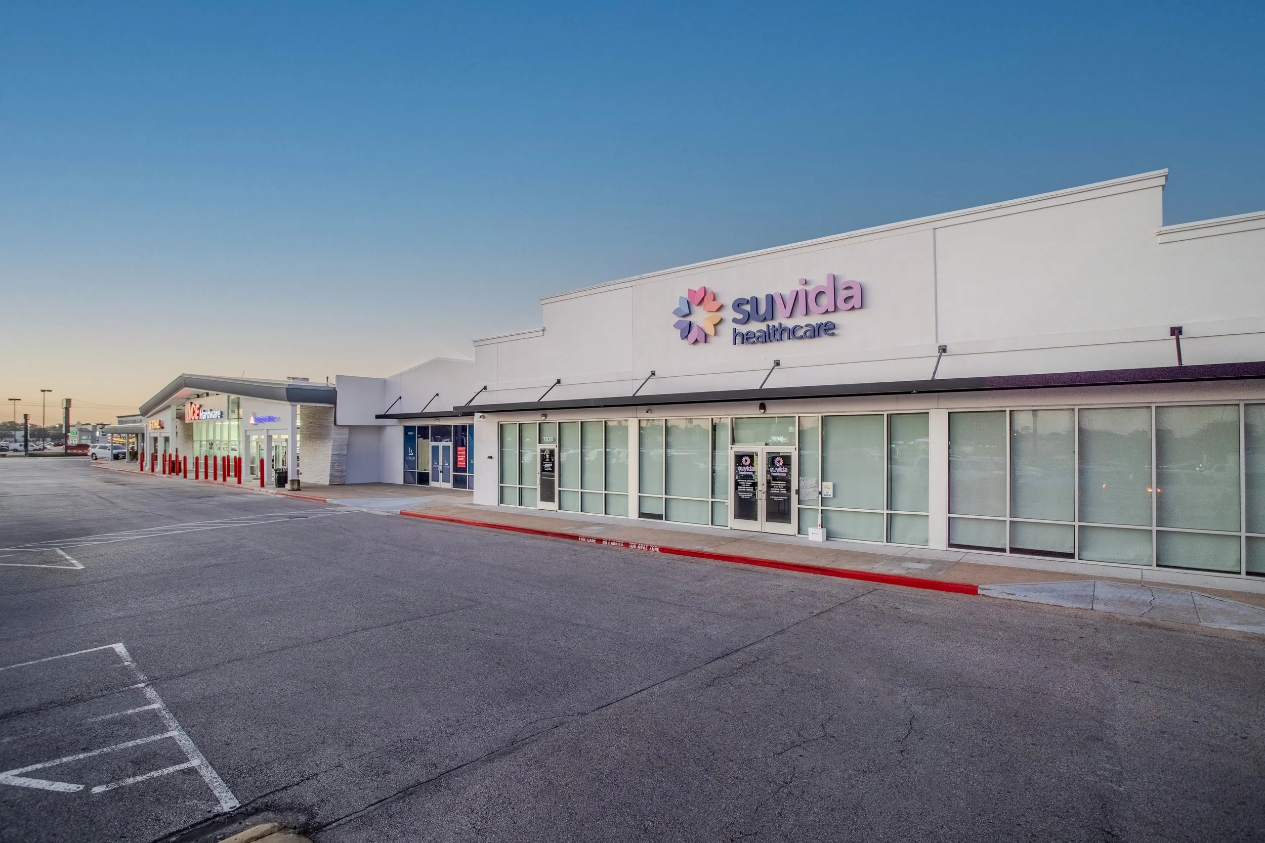 Empty shopping plaza with Suvida Healthcare store in the foreground and other stores in the background, parking lot with marked spaces, and a clear sky.