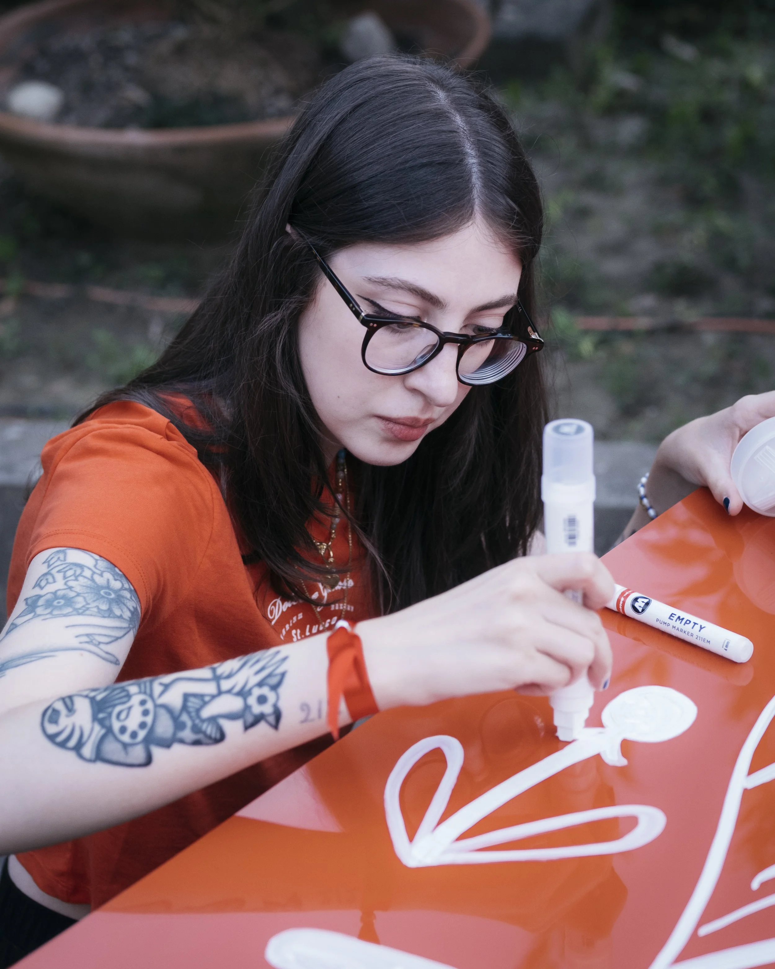 Una giovane donna con tatuaggi sulle braccia, occhiali e maglietta arancione disegna su una superficie di colore arancione con un pennarello bianco. Sta concentrata mentre crea un disegno.