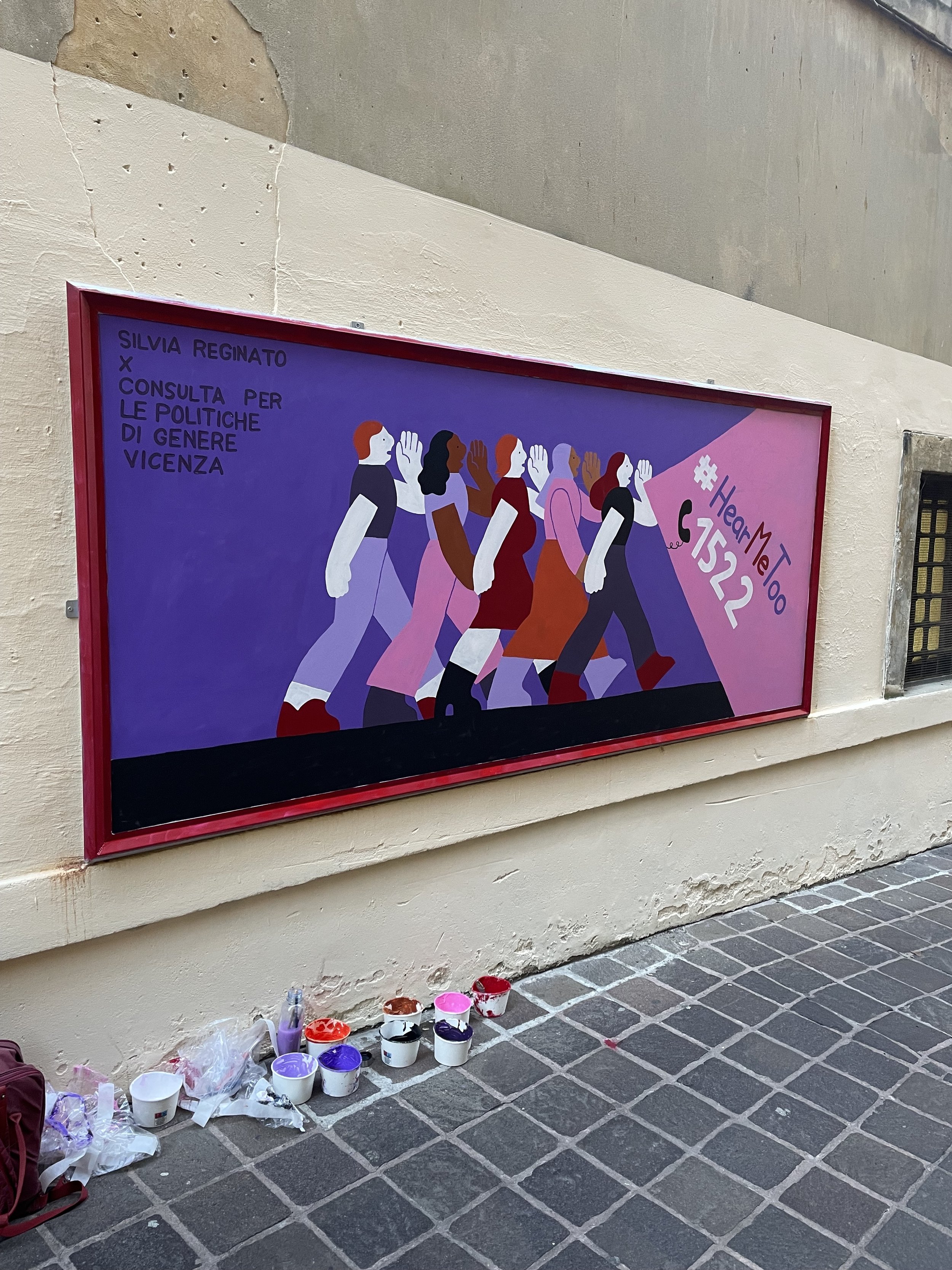 Murale colorata di donne di diverse etnie che camminano in fila. I colori principali sono viola, rosa, rosso e nero. Appena a destra, frase #HearMeToo 1522 e icona di un telefono