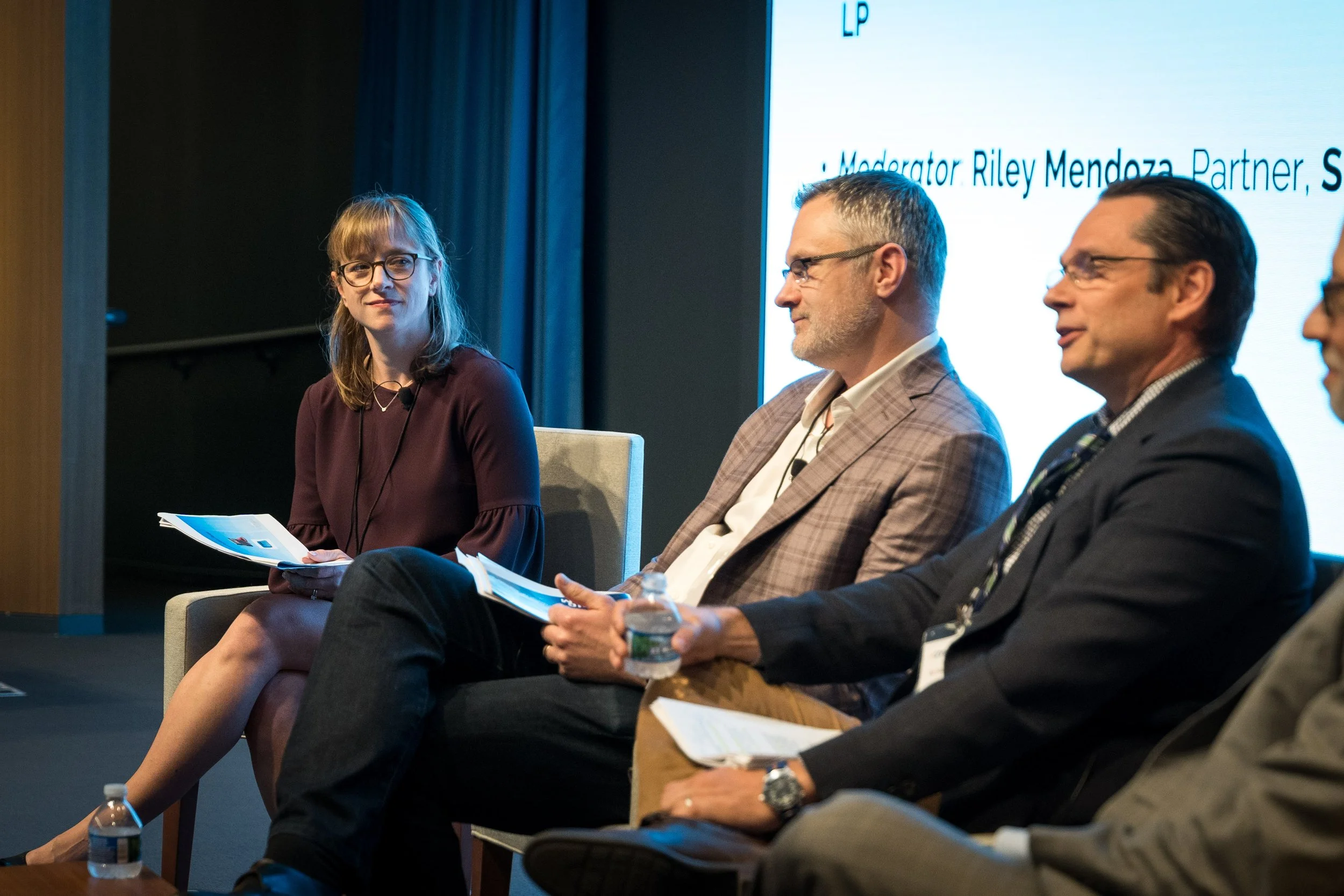 Panel of four people sitting on stage at a conference, with one woman on the left holding papers and three men on the right. Behind them is a large screen with text.
