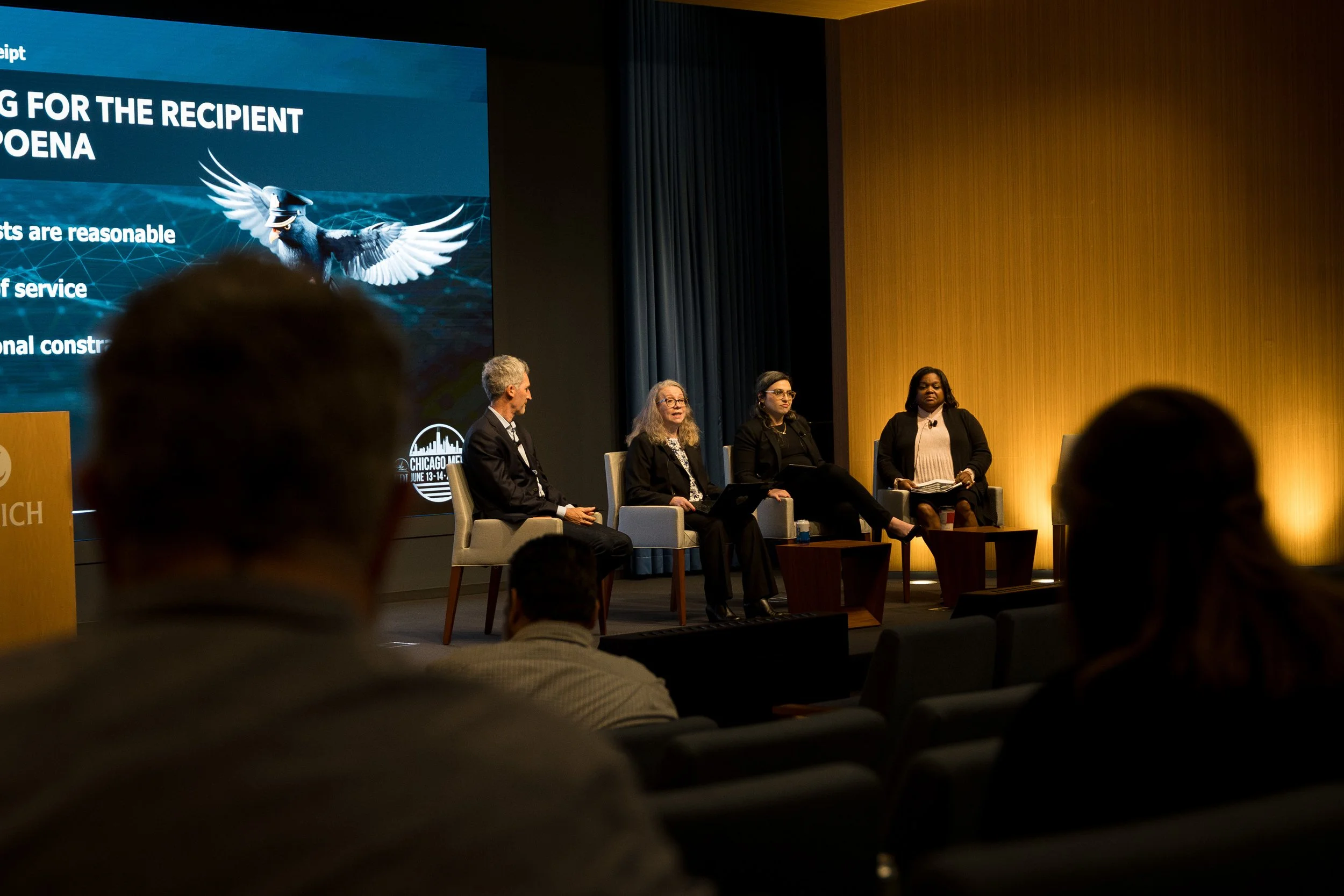 A panel discussion at a conference with four women and one man seated on stage, with a large screen behind them displaying a graphic of a bird with a graduation cap and partial text about criteria for the recipient.