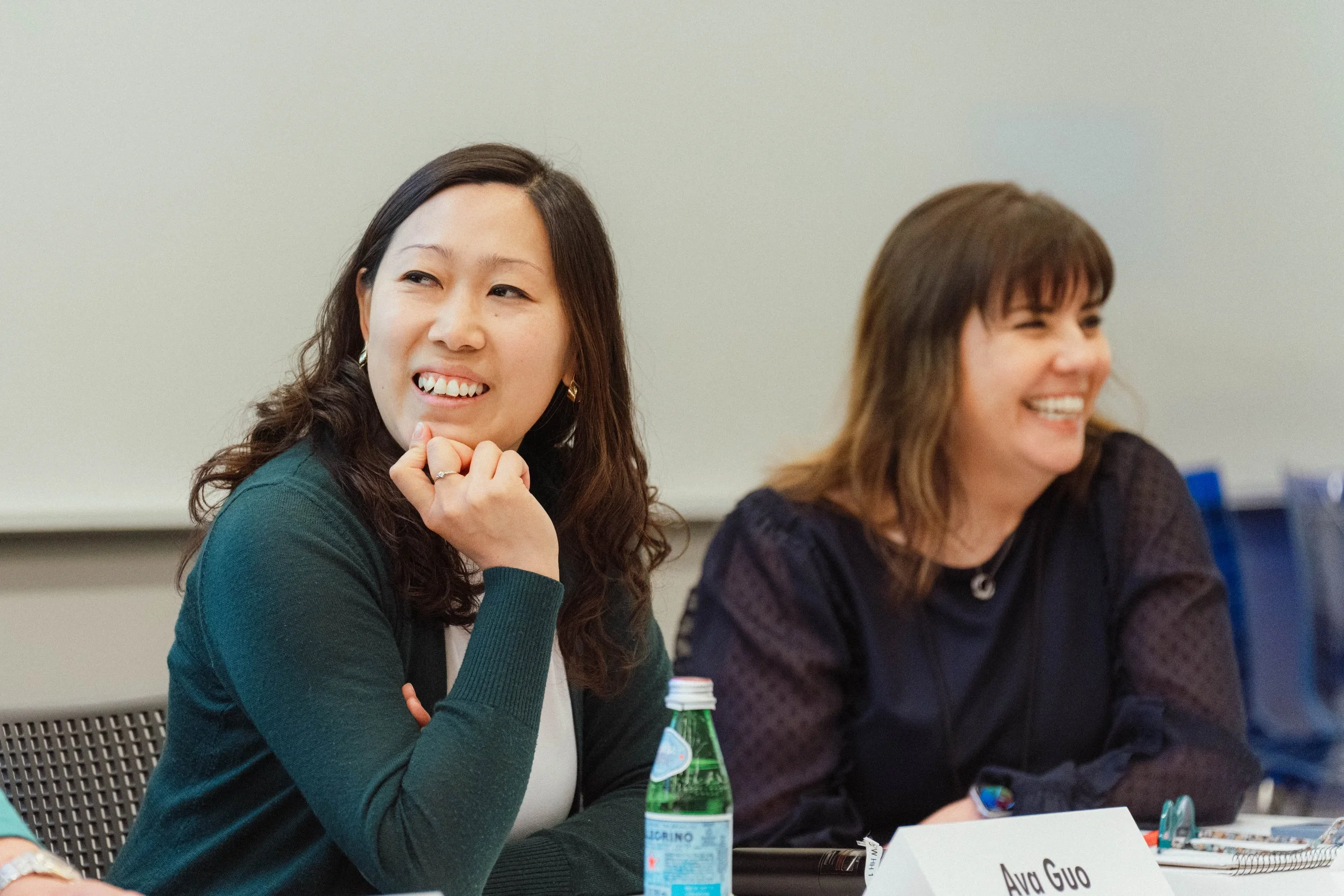 Two women sitting at a table, smiling and engaged in a discussion. One woman has dark hair, wearing a green jacket, and the other has brown hair with bangs, wearing a black top. A water bottle and name tags are visible.