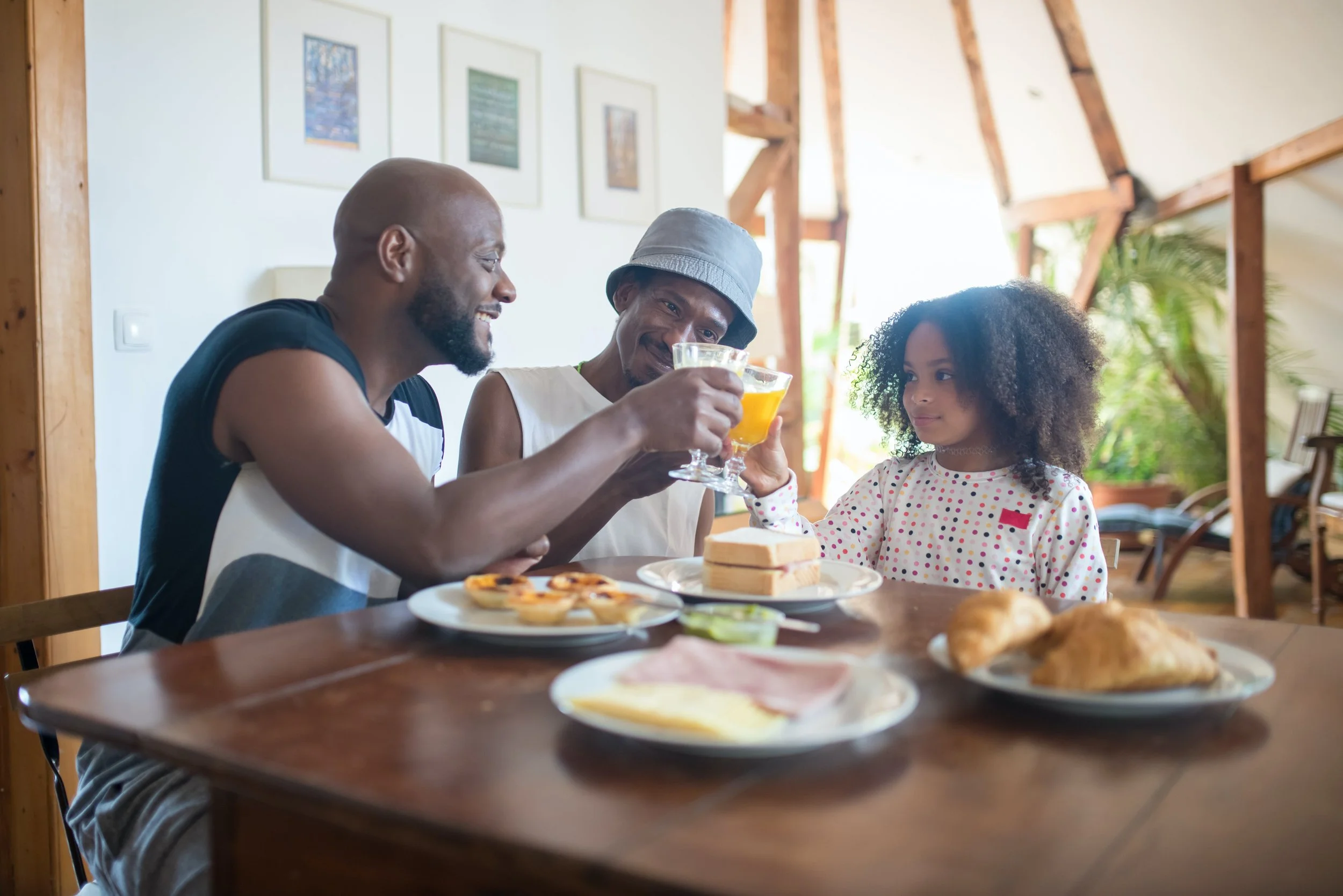 A happy Black gay male couple sitting at a kitchen table smiling with their young daughter, representing inclusive LGBTQ+ affirming family therapy at Neutral Ground Counseling Group in Fairfax, VA.