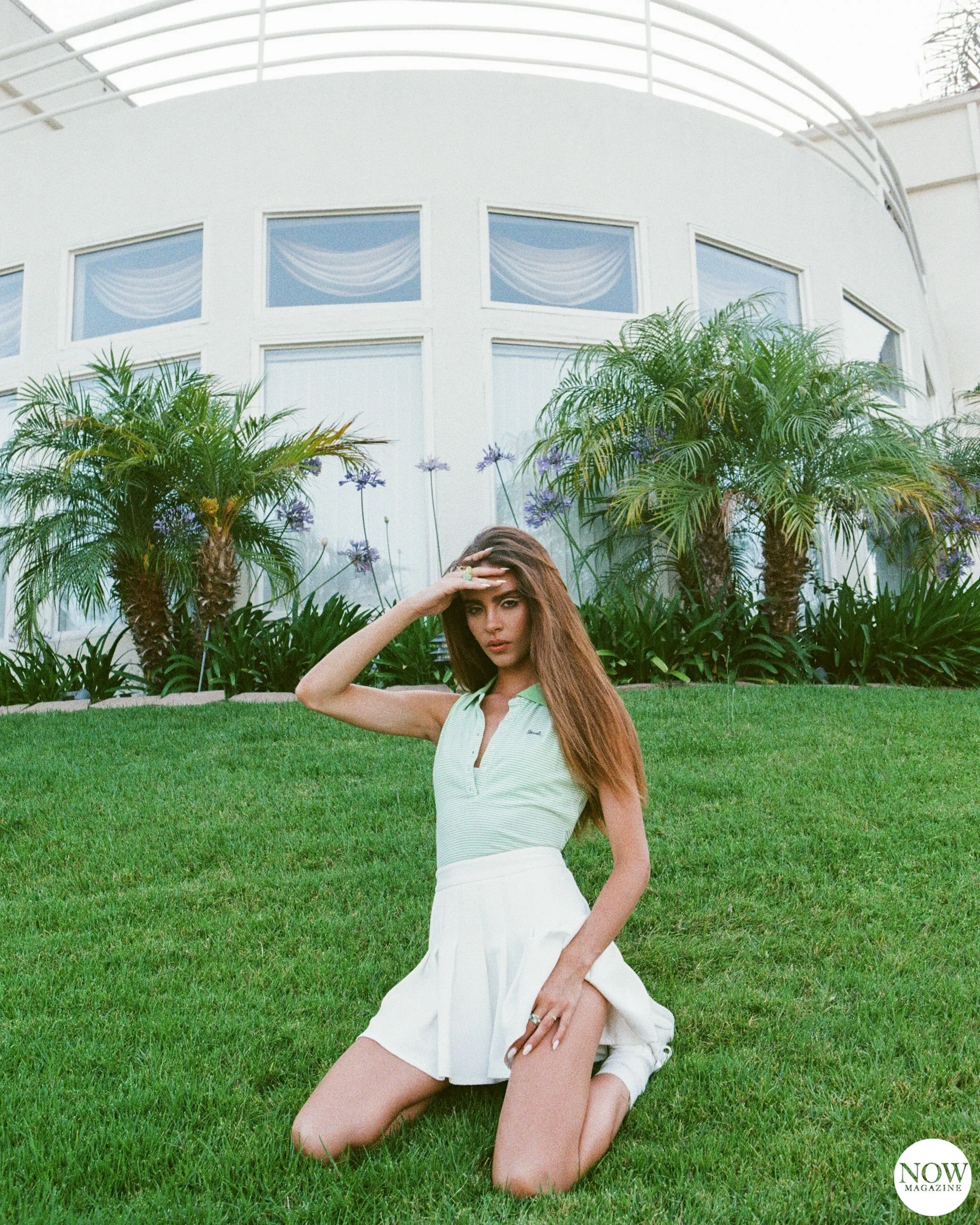 A woman kneeling on grass in front of a white building, wearing a green sleeveless top and a white pleated skirt. She is posing with one hand shielding her eyes from the sunlight. Palm trees and plants are visible in the background.