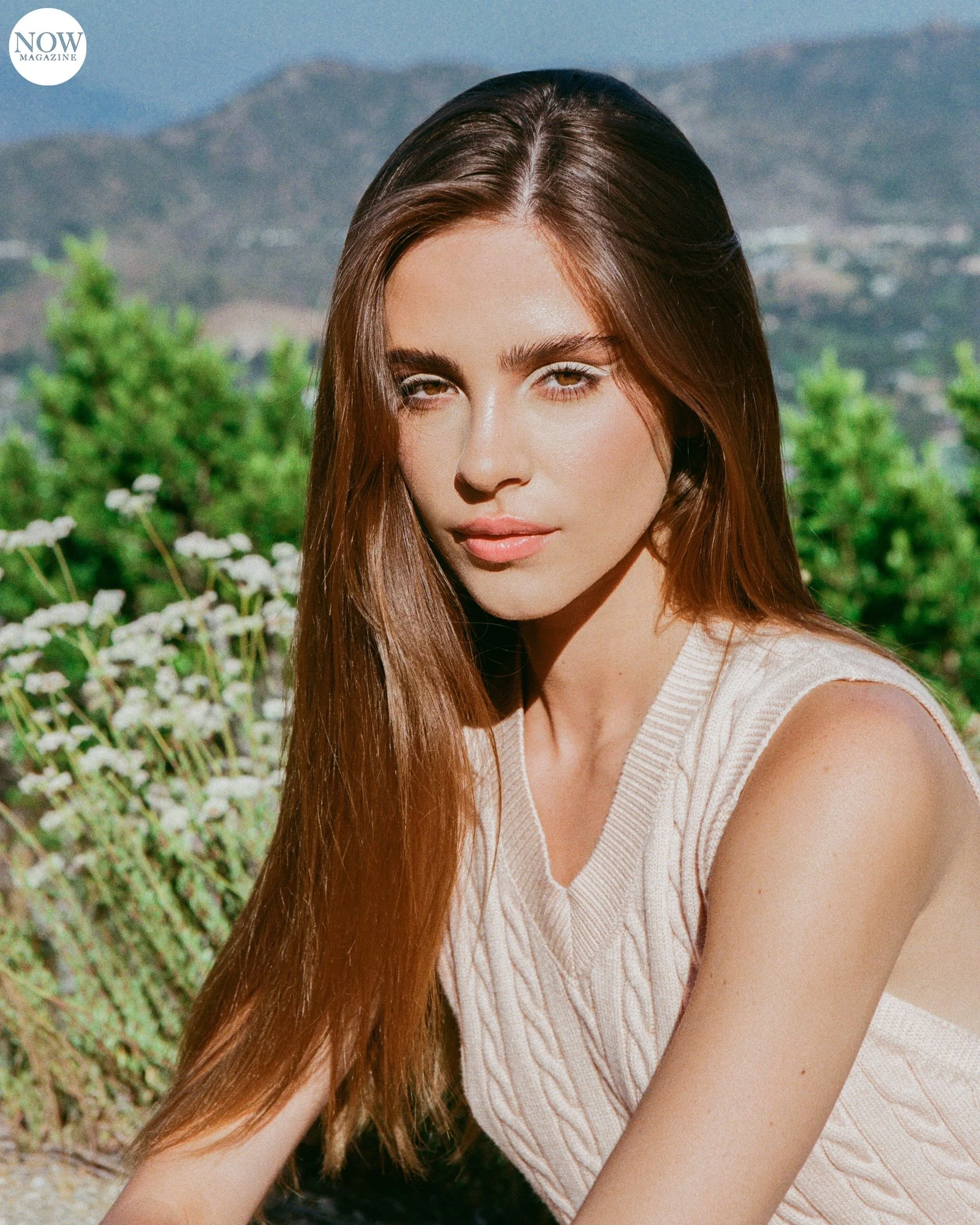 Woman in a sleeveless knit top with long brown hair, outdoors with greenery and mountains in the background.