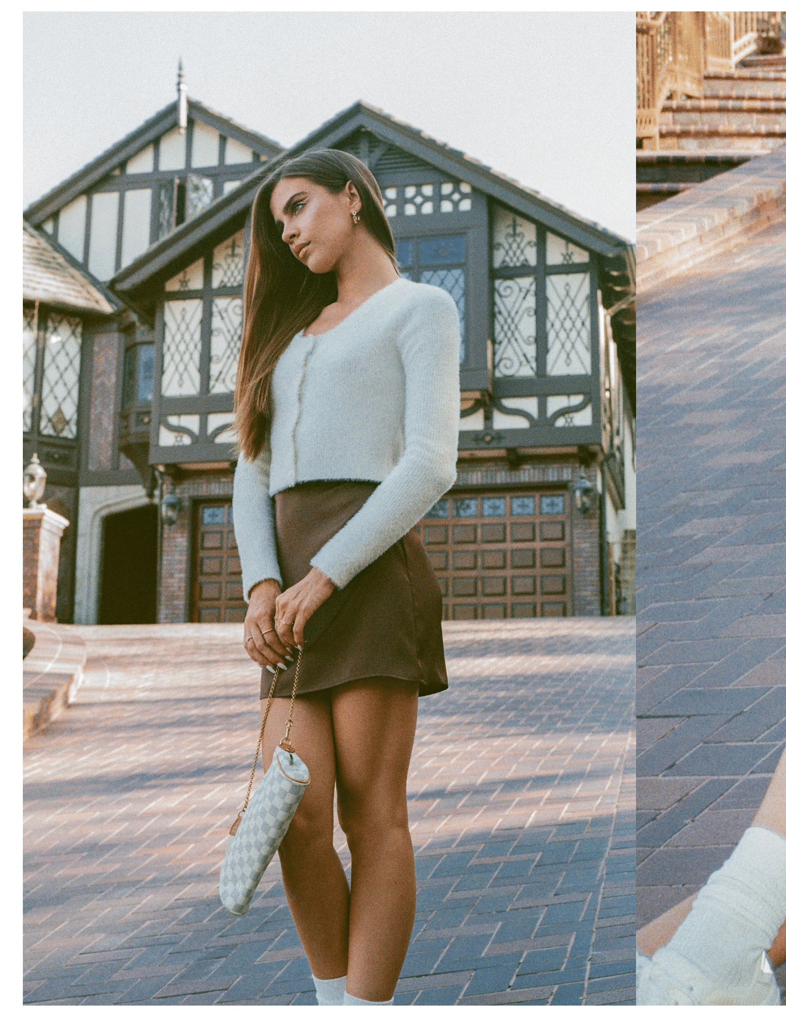 Woman in elegant outfit standing on brick driveway with historic-style house, holding a patterned handbag, wearing a light cardigan and brown skirt.