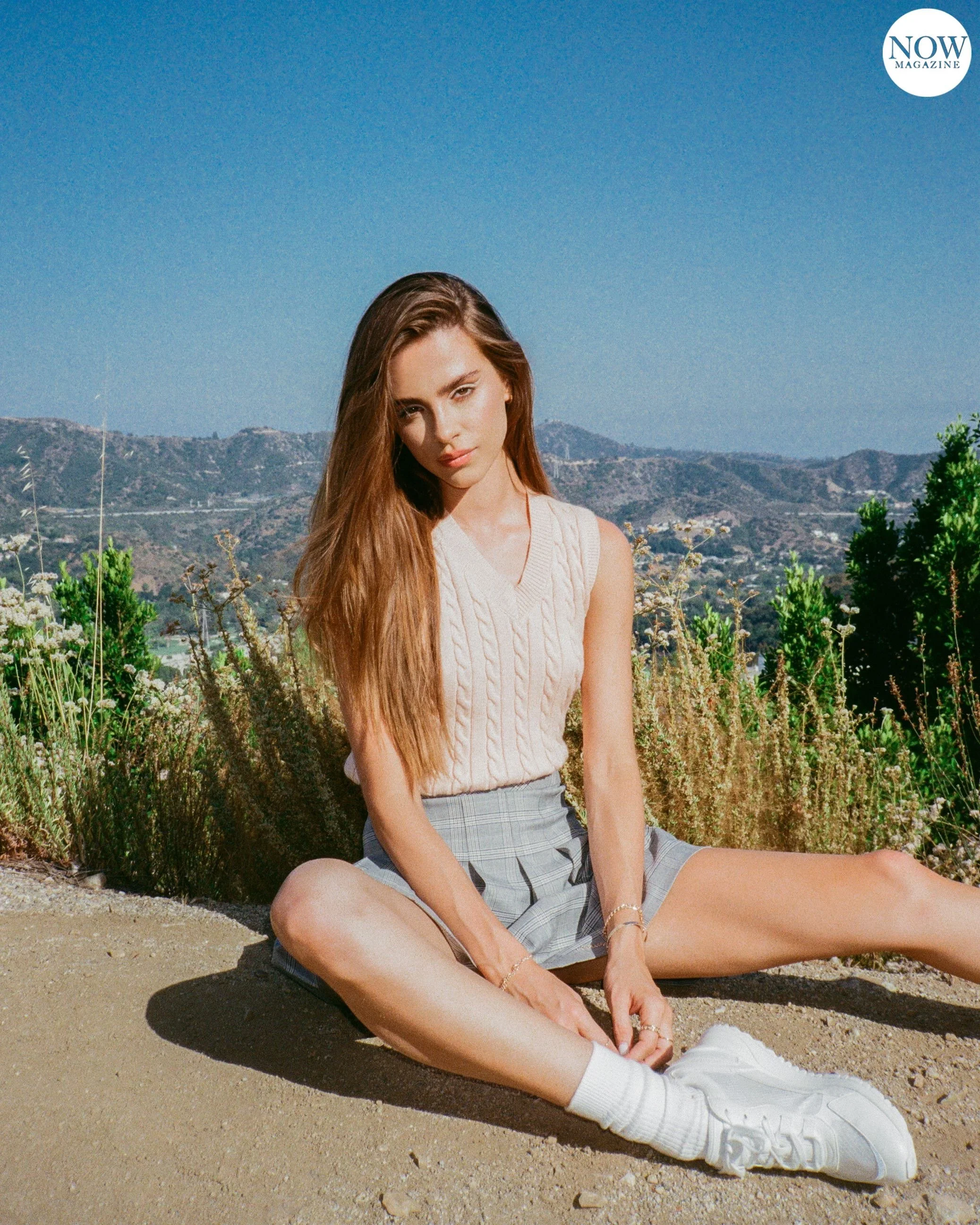 Young woman in a sleeveless top and plaid skirt sitting on the ground outdoors with a scenic mountain view in the background.