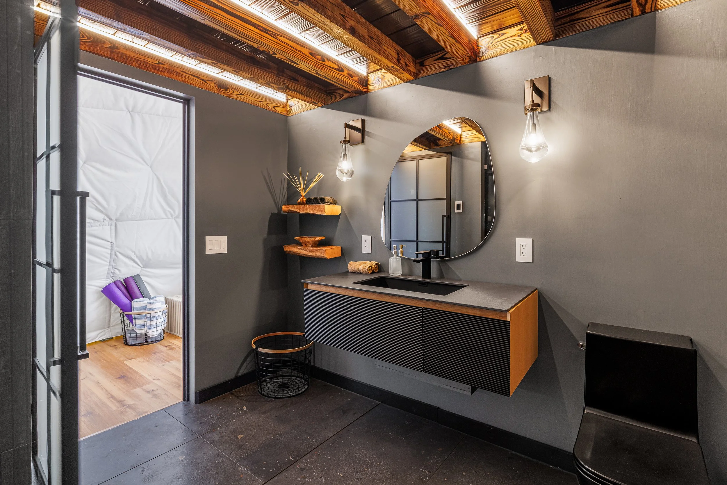 Modern bathroom with a gray wall, black vanity with wood accents, oval mirror, and two wall-mounted lights with exposed bulbs.