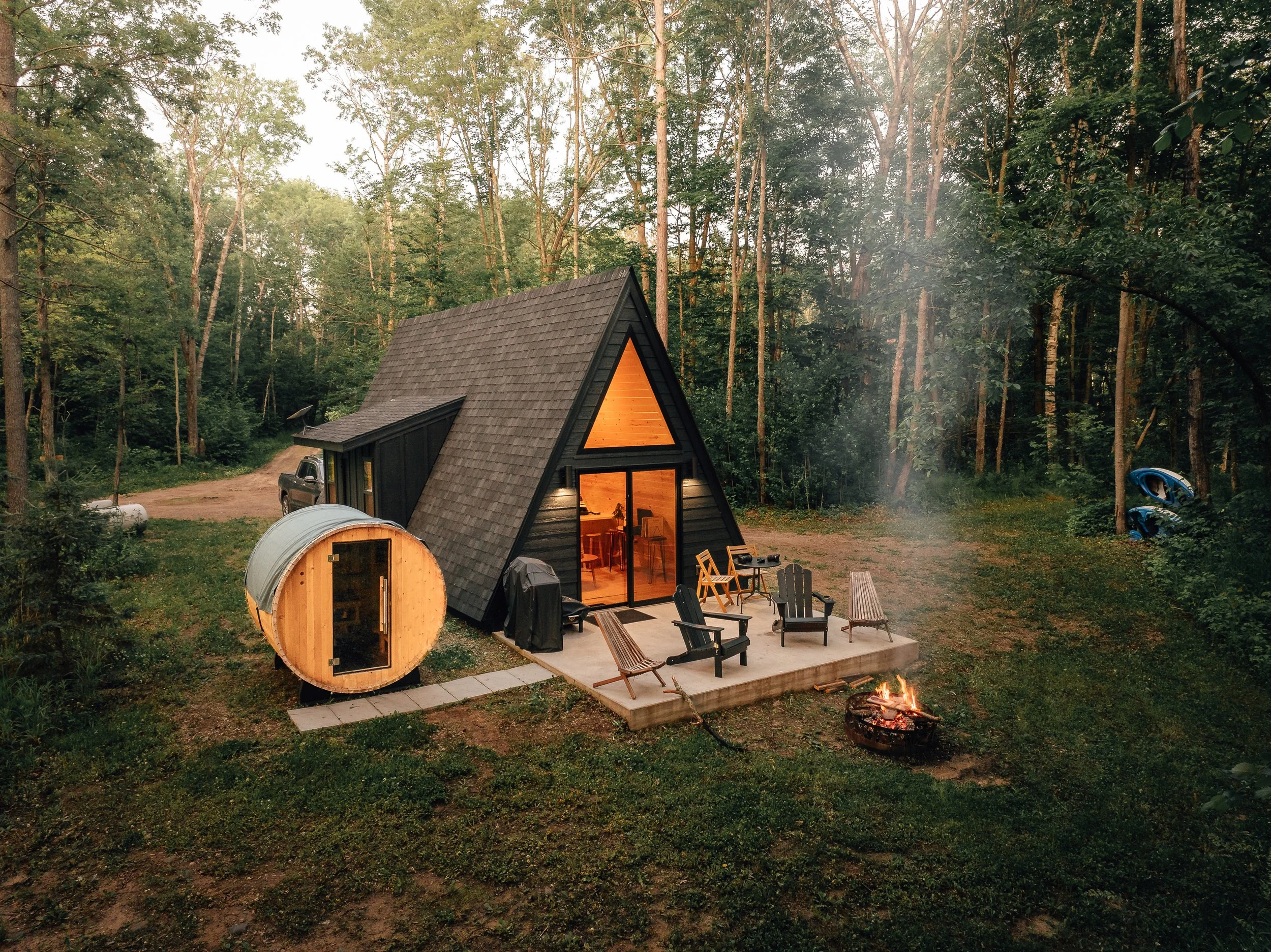 A small black A-frame cabin with a wooden interior visible through sliding glass doors, situated in a forest. A wooden deck with chairs and a fire pit is outside. A metal barrel sauna is nearby, and kayaks are stacked to the right. The scene is warm