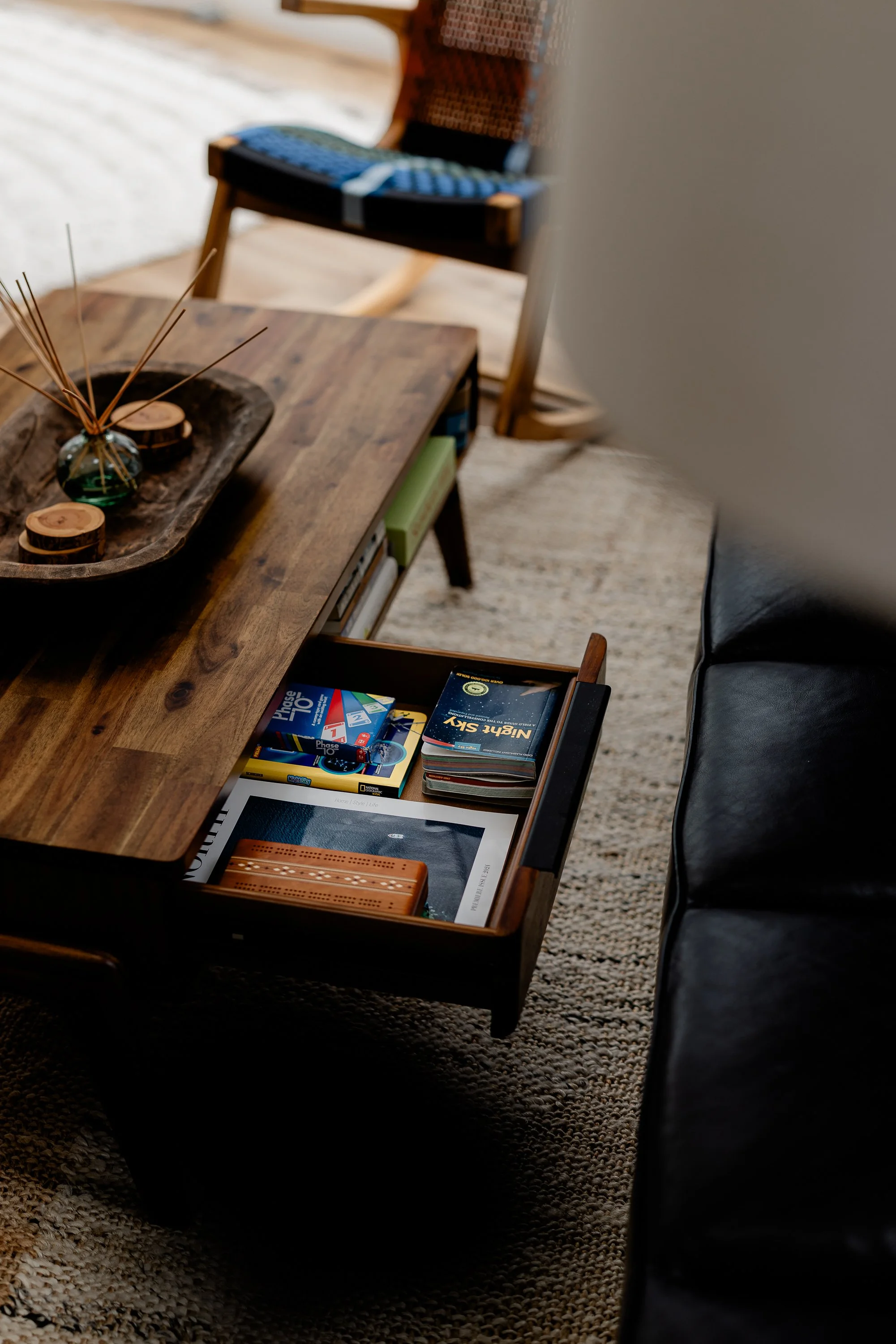 A wooden coffee table with various items including a reed diffuser, candles, and books, partially pulled out a drawer containing more books and a framed photo, situated in a cozy living room with a black leather sofa and a woven chair in the backgrou