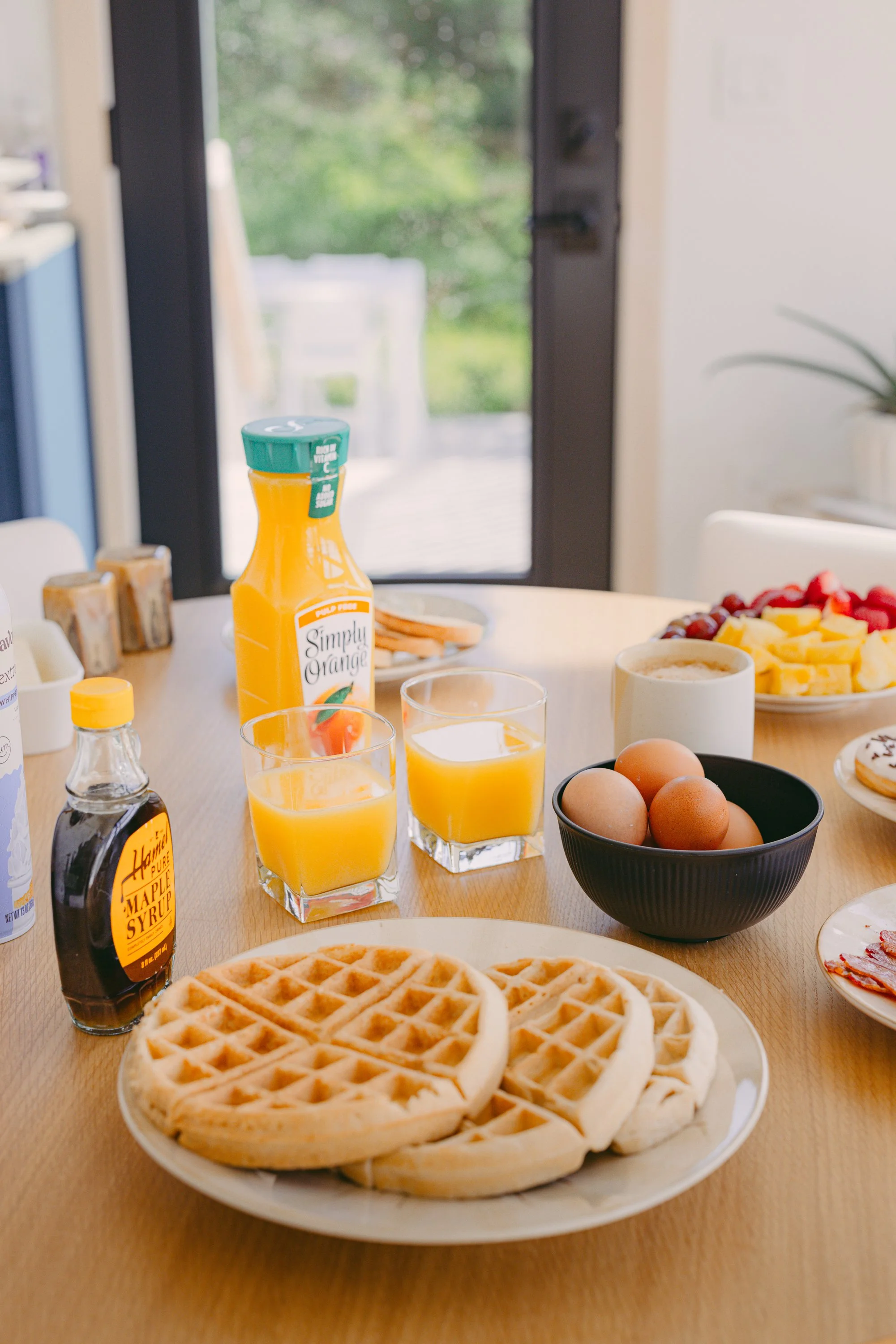 A breakfast spread with waffles, eggs, orange juice, syrup, and fruit on a wooden table near a sliding glass door.