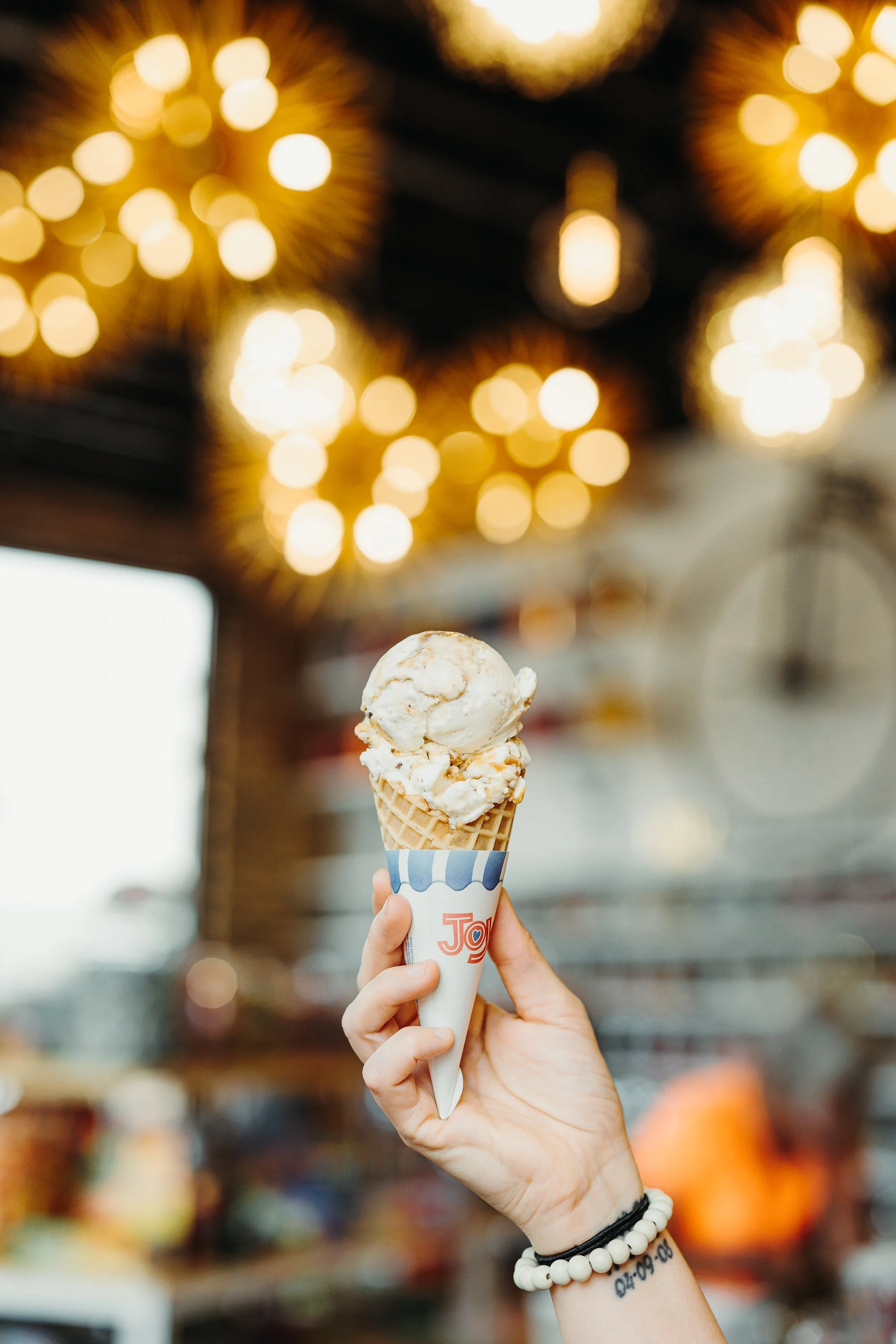 A hand holding a vanilla ice cream cone with caramel topping in a shop, with warm yellow lights creating a bokeh background.
