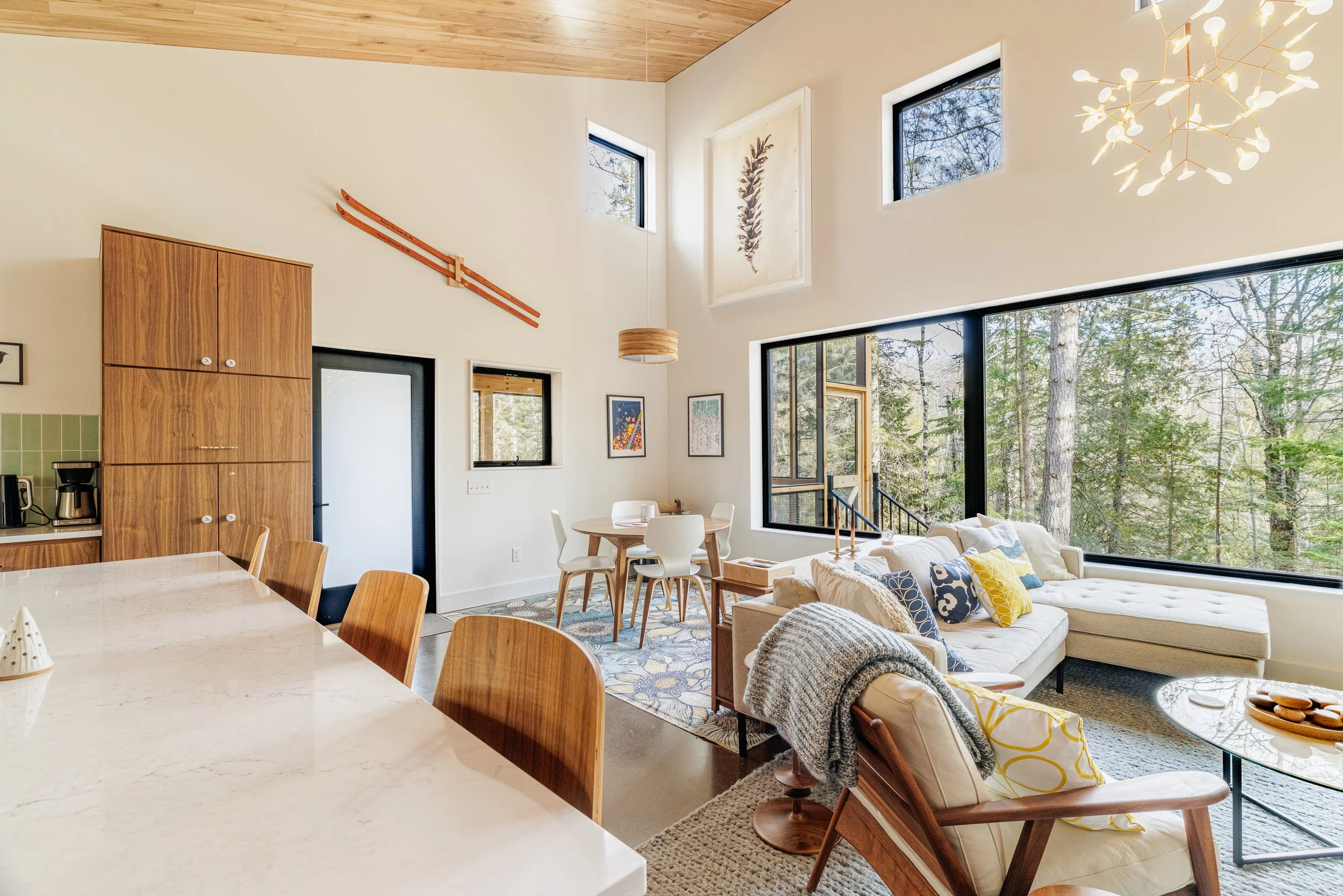 Open concept living and dining area with large windows, white walls, wooden ceiling, modern furniture, decorative art, and forest view