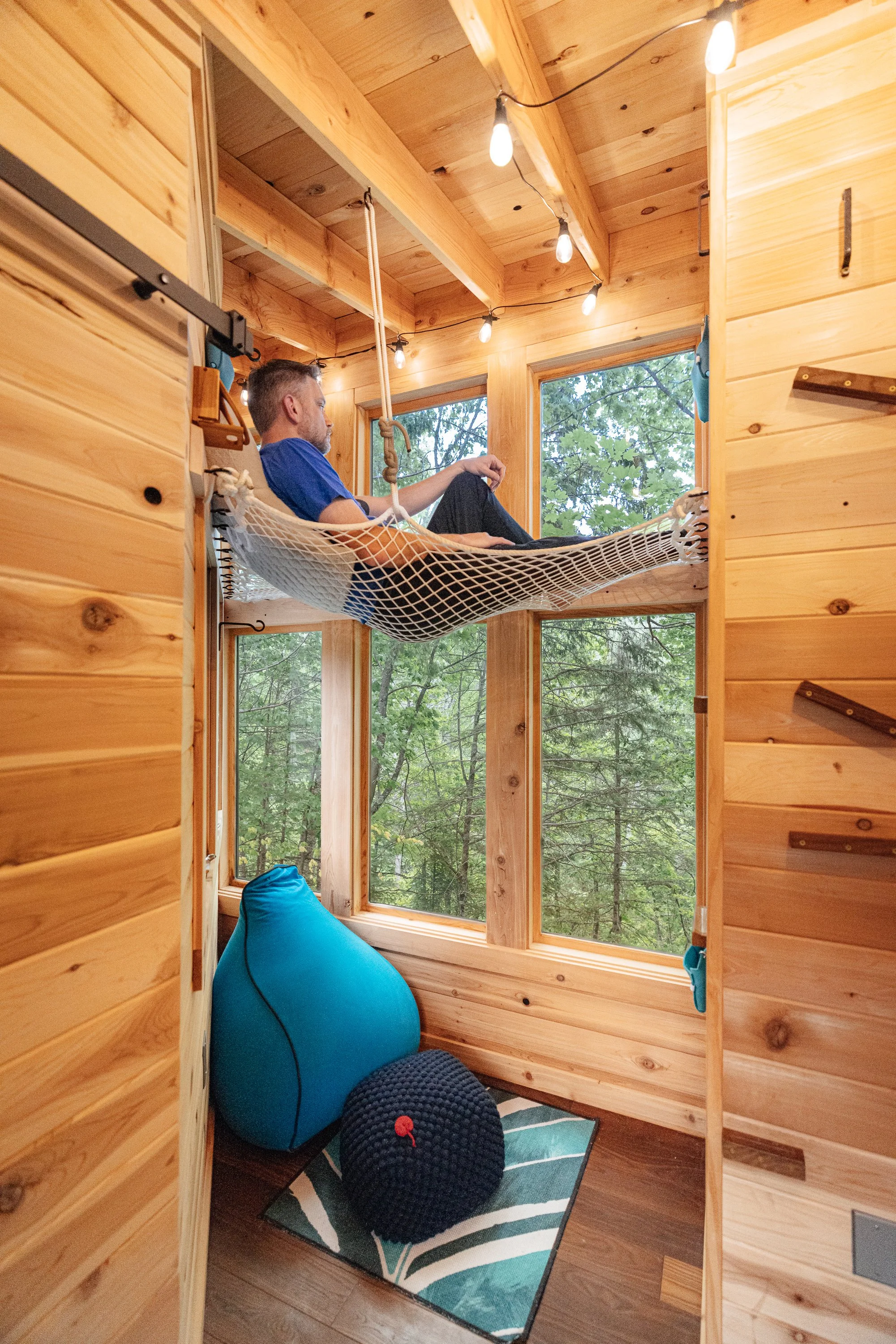 A person relaxing in a hammock inside a wooden cabin with large windows showing green trees outside.