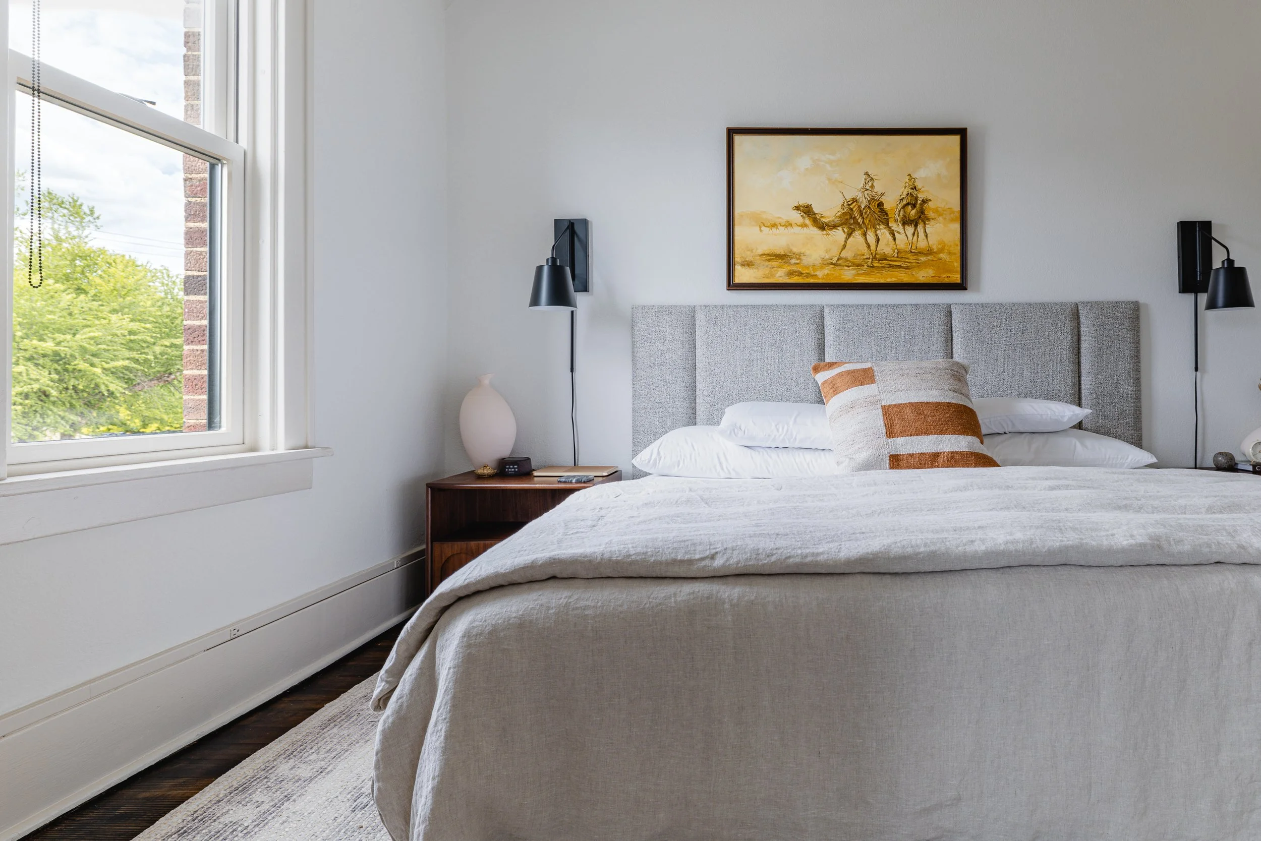 Clean and cozy bedroom with a large bed, white bedding, a striped pillow, nightstand with a lamp, vase, and phone, window with greenery outside, and a framed painting of cowboys on horseback above the bed.