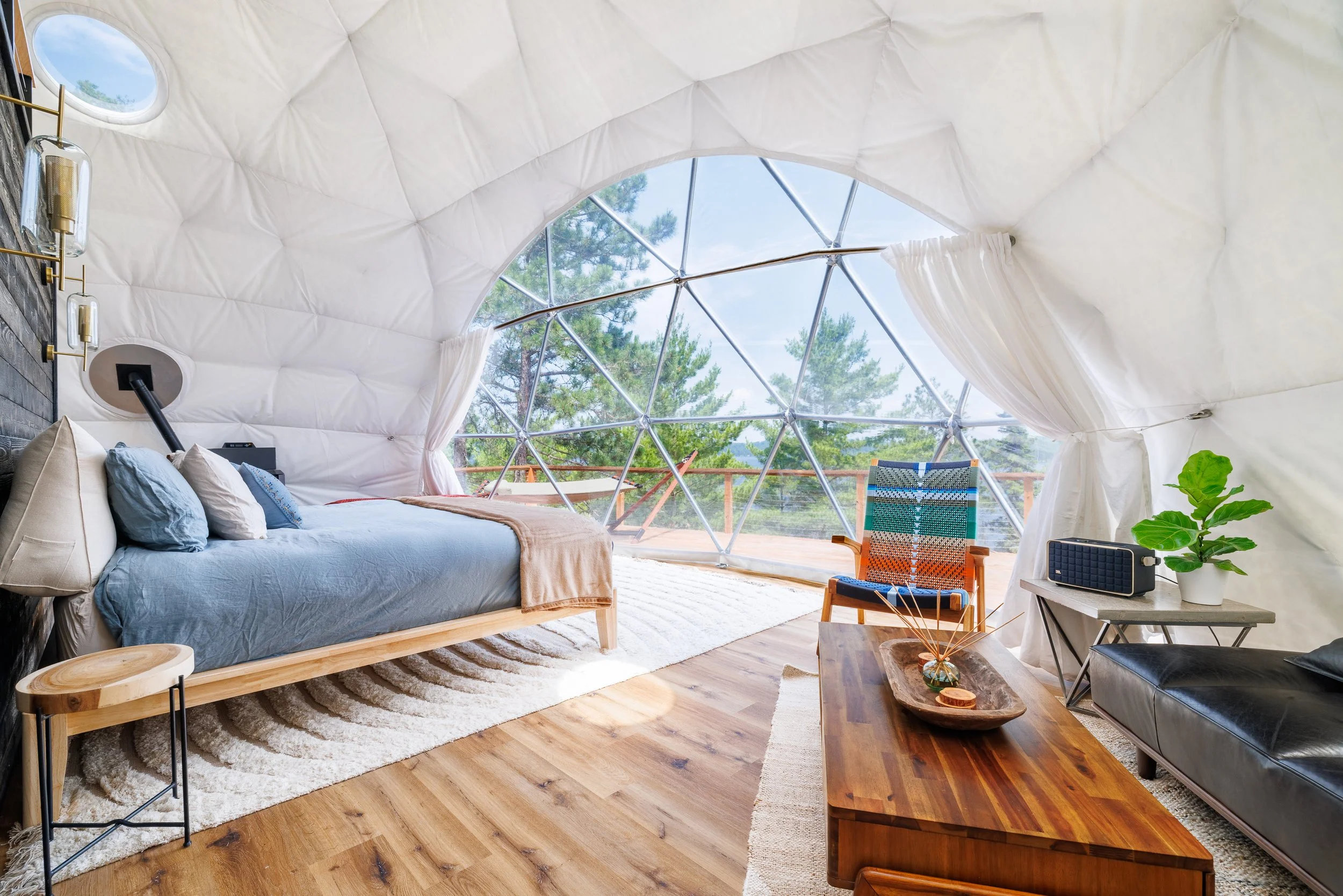 Interior of a modern geodesic dome bedroom with a bed, armchair, coffee table, and large windows overlooking trees.
