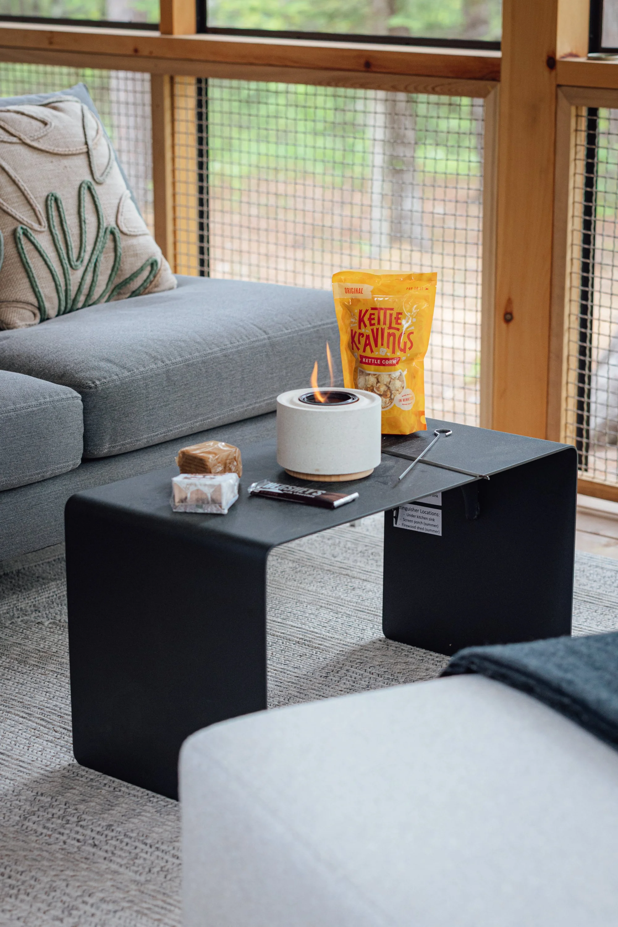 Living room with a black coffee table holding a bag of kettle corn, two wrapped snacks, a chocolate bar, and a small ceramic burner with a flame. There is a gray couch with a decorative pillow and a wooden-framed screen door in the background.