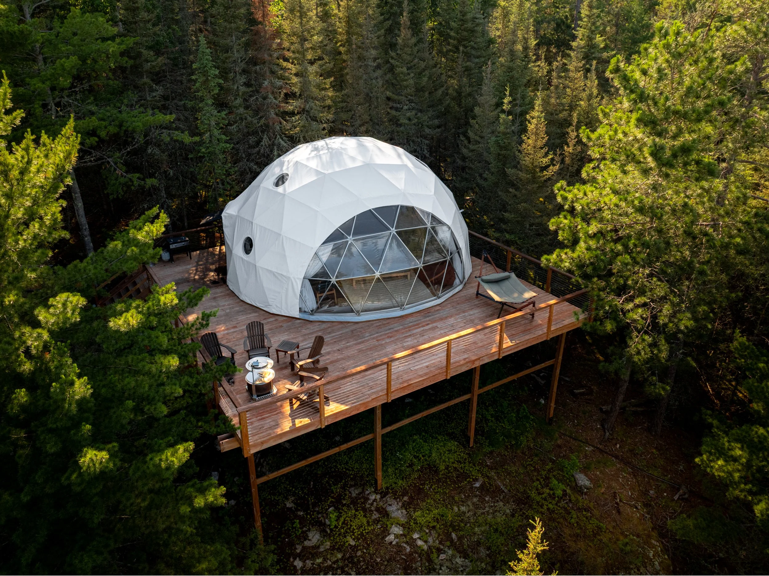 An outdoor deck with a geodesic dome tent in a forested area, furnished with outdoor chairs, a fire pit, and a hammock.