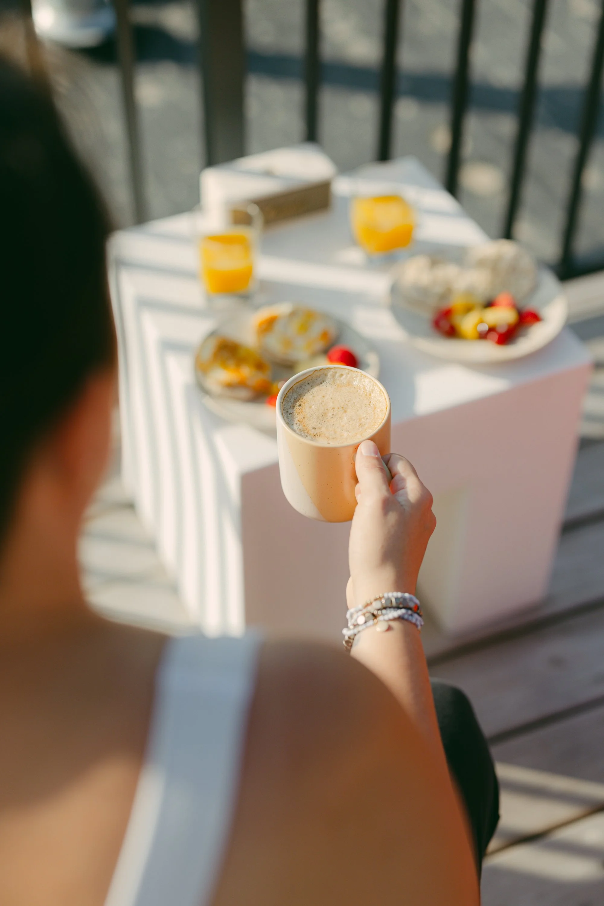 A woman holding a coffee mug on a balcony table with breakfast items such as orange juice, muffins, and fruit.